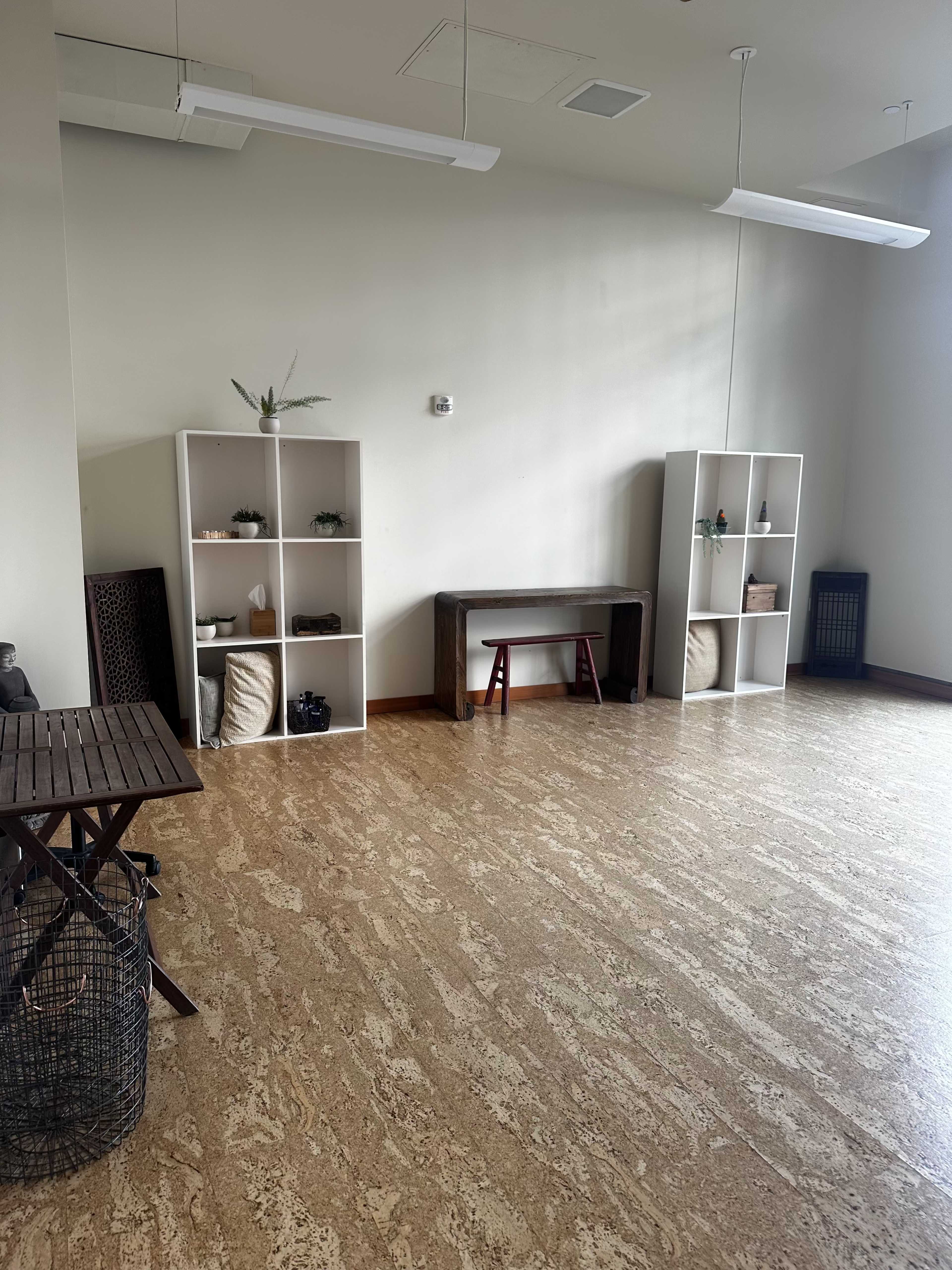 A spacious, minimally furnished room with two white shelving units, a wooden bench, a small table, and a wicker basket on the floor.