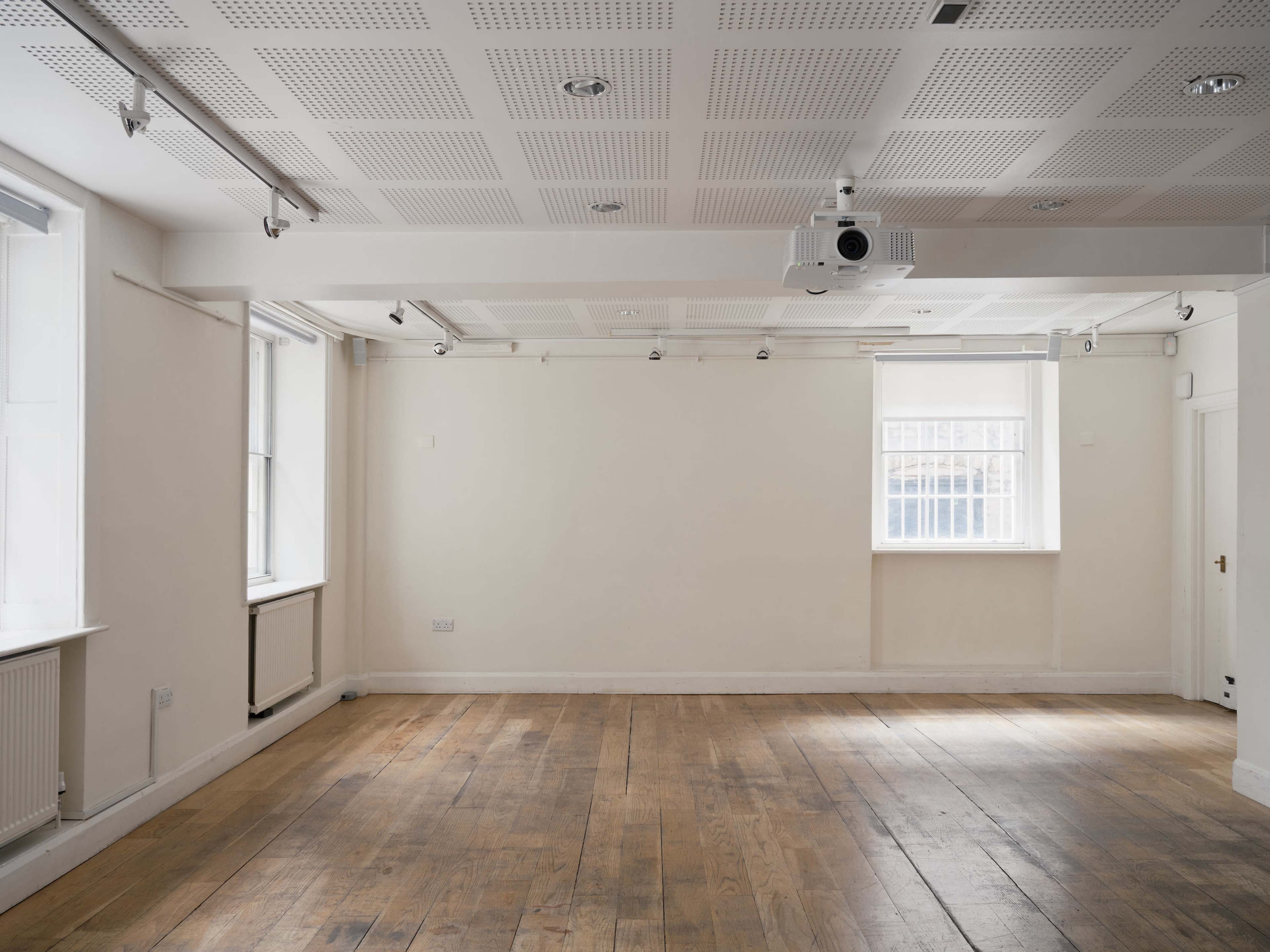 The image shows an empty room with wooden flooring, white walls, and a ceiling-mounted projector.