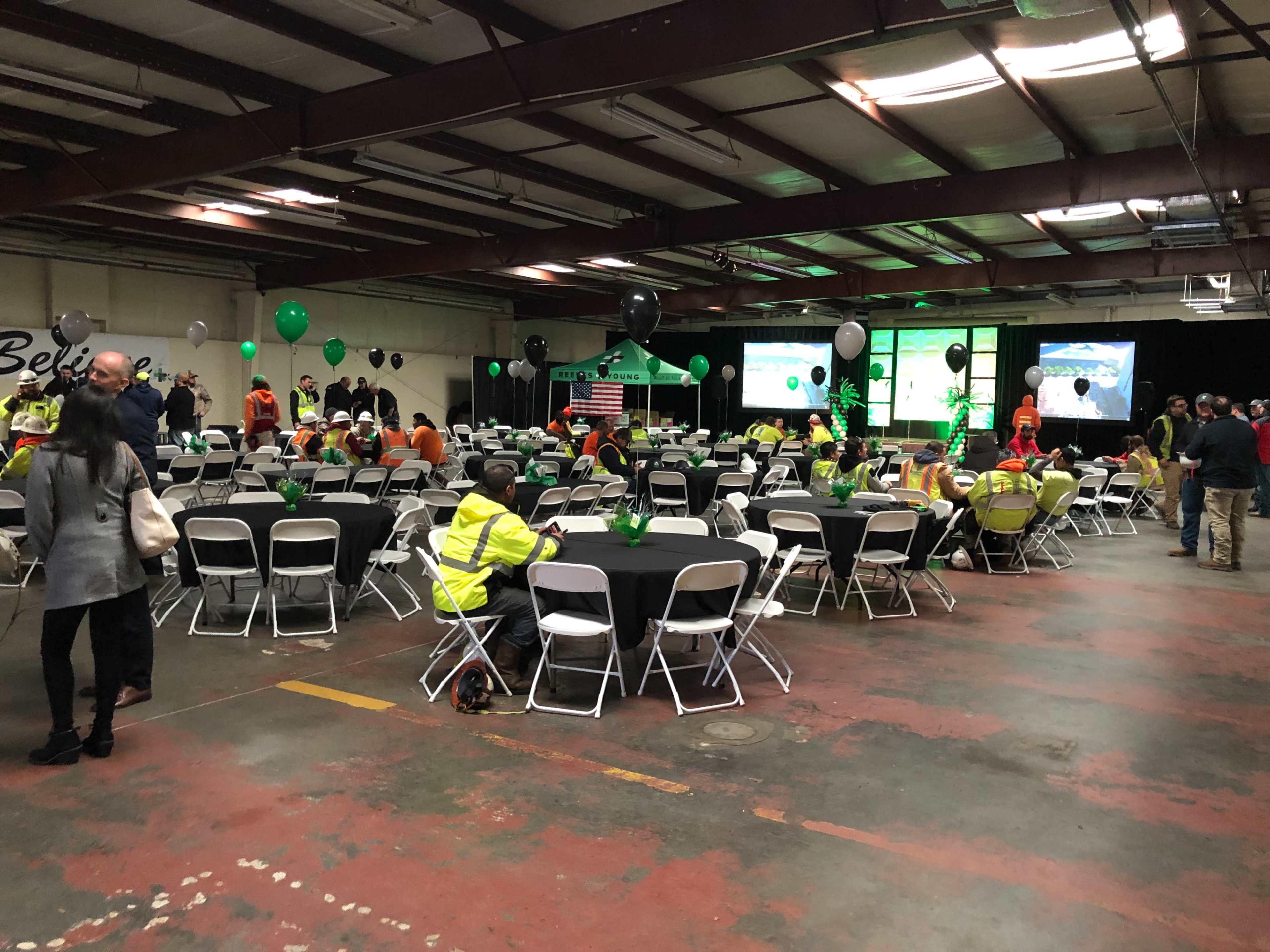 The image shows a large indoor space set up for an event, featuring a series of tables with chairs, decorations including balloons, and screens displaying presentations, with people seated and mingling.