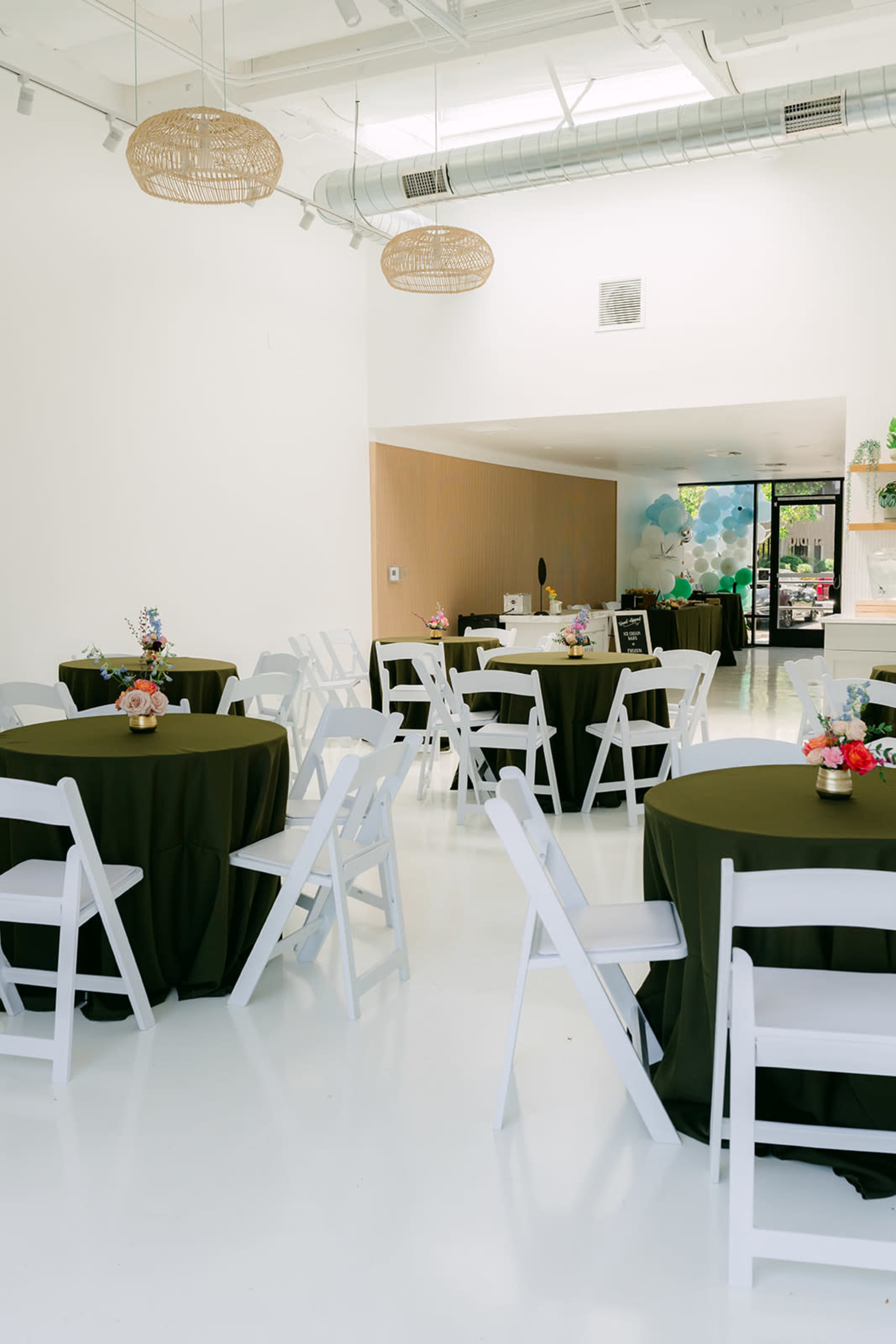 The image shows a spacious, bright room with round tables covered in dark green tablecloths, white folding chairs, and floral centerpieces, along with a counter area at the back.