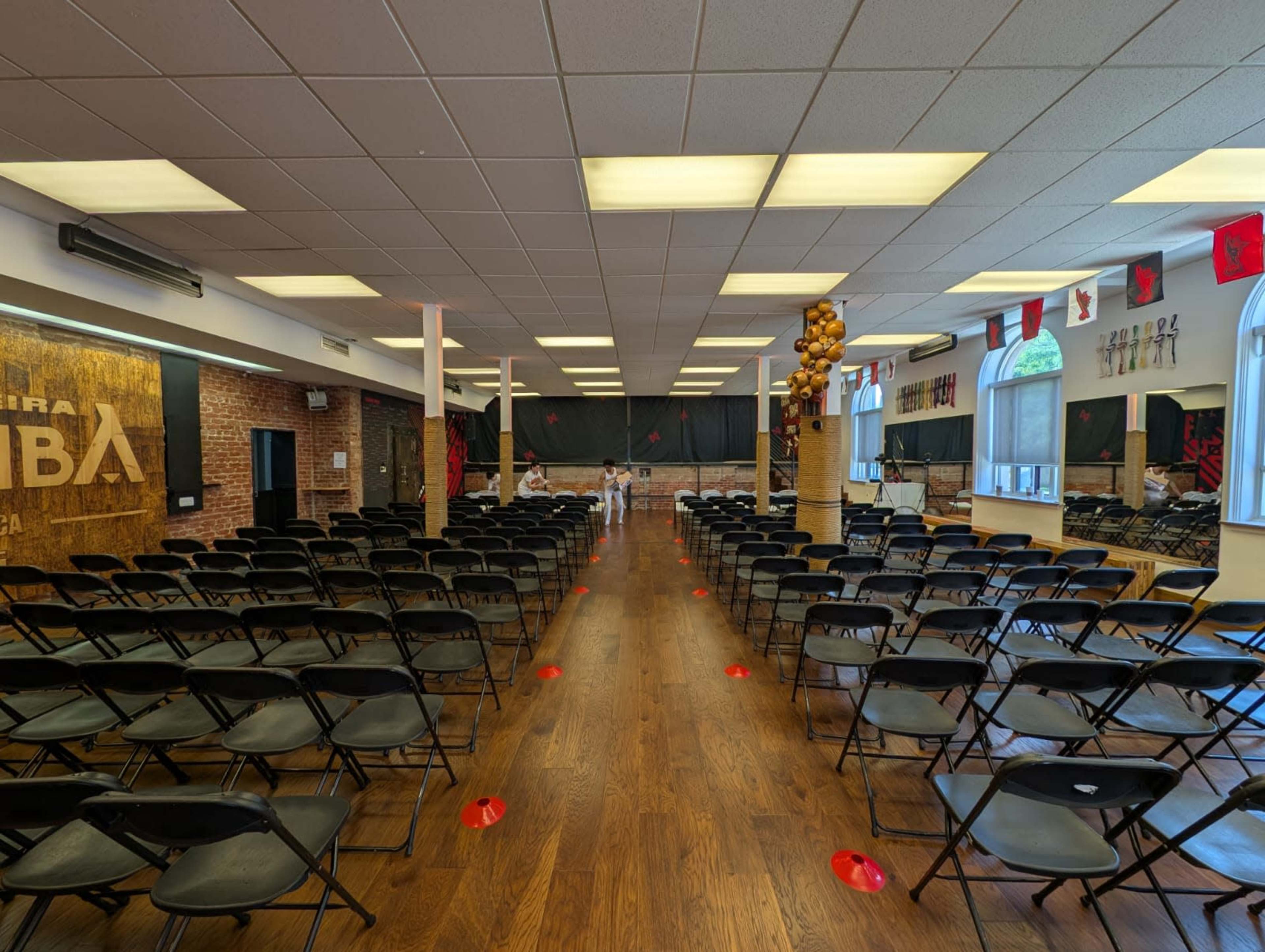 The image shows a large room set up with rows of black chairs facing a stage, with red markers on the floor and a brick wall in the background.