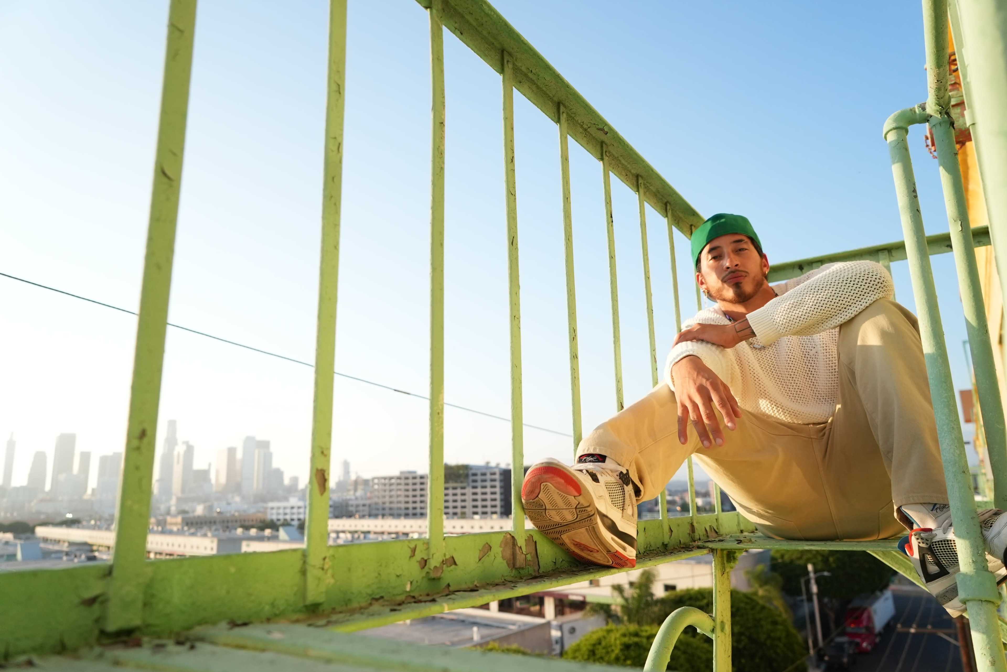 A man sits on a green railing overlooking a city skyline during sunset.