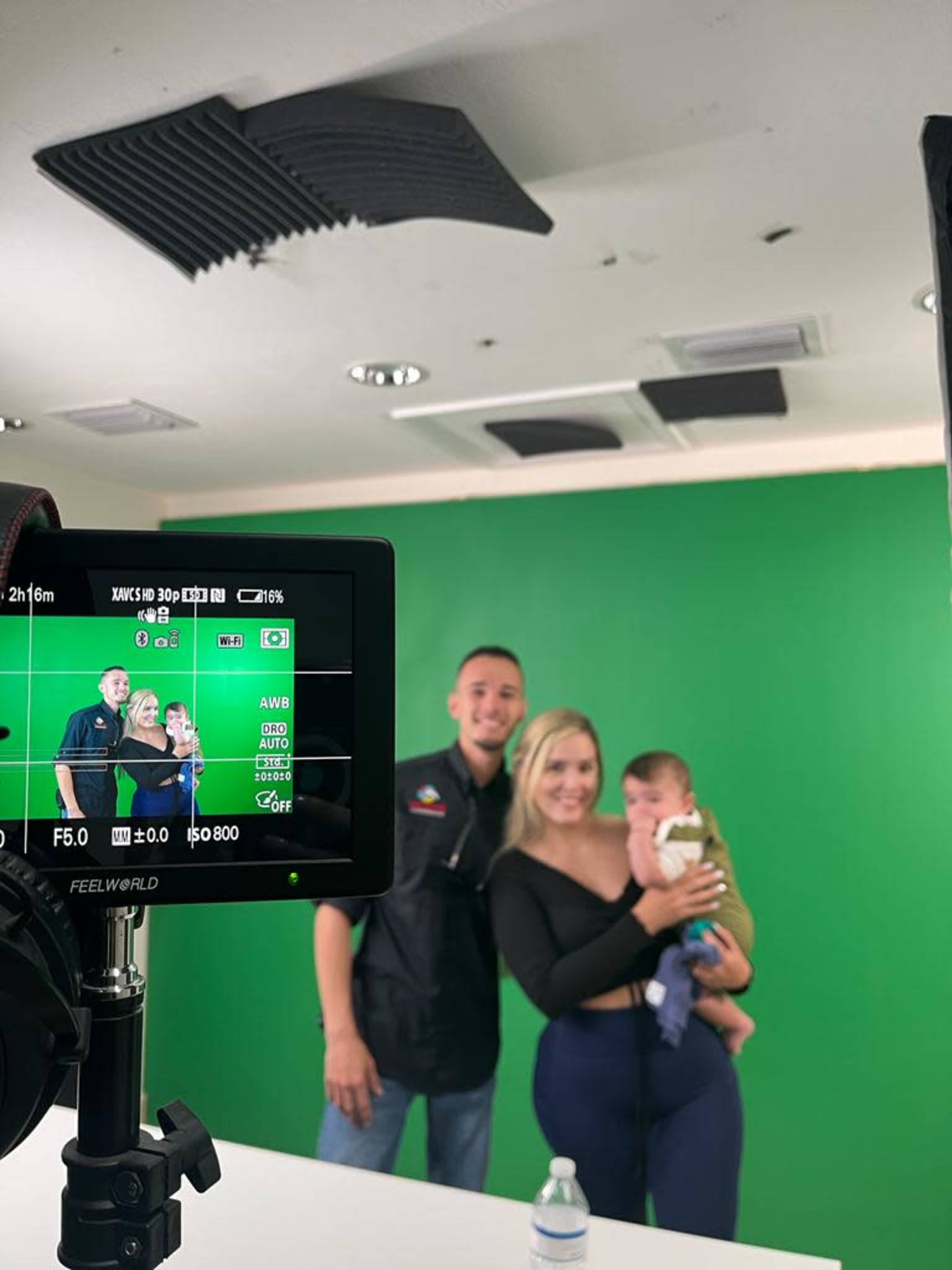 A couple poses with a baby in front of a green screen while a camera screen displays their image.