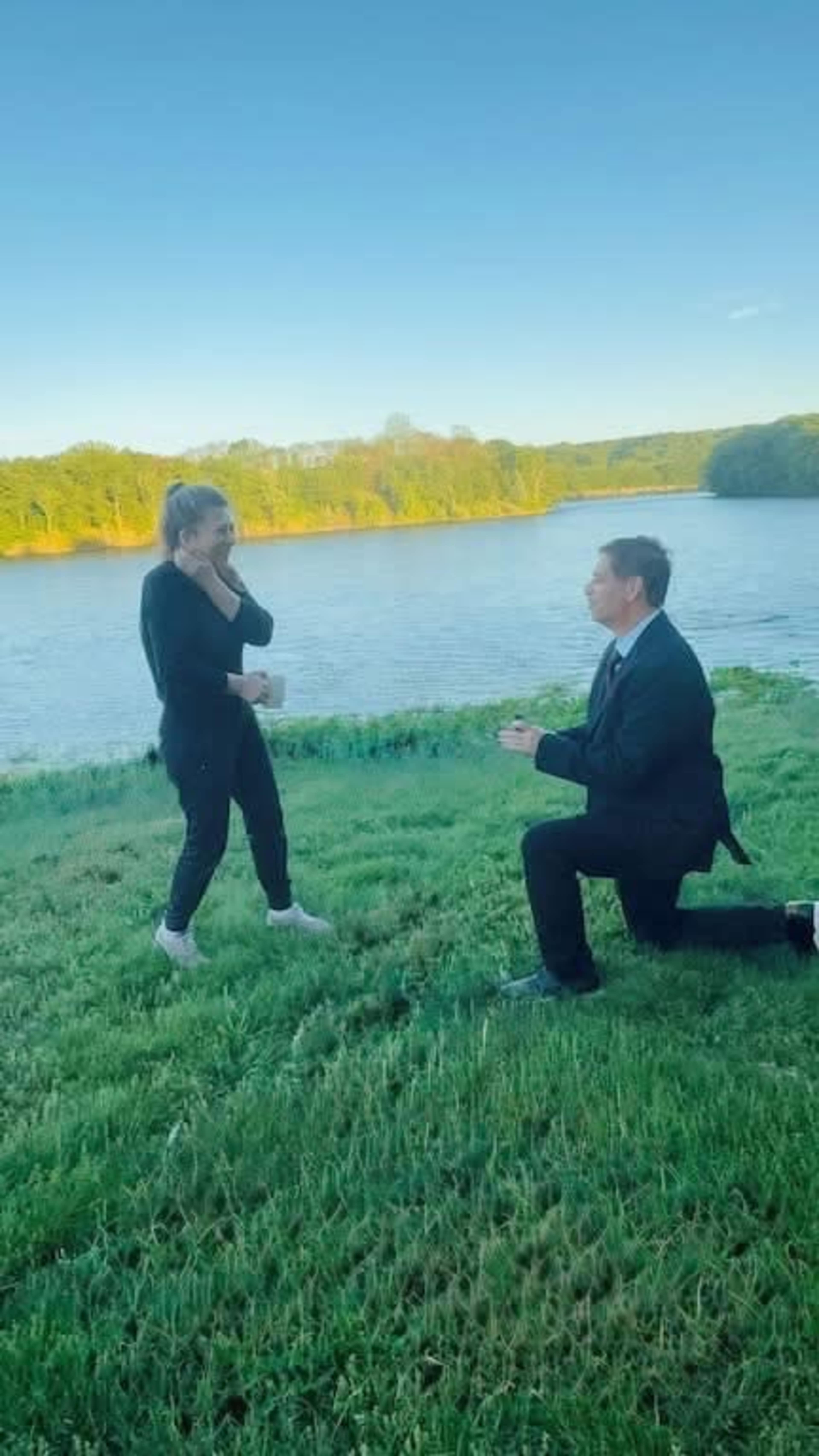 A man in a suit kneels on one knee in front of a woman by a river, proposing while she appears surprised.