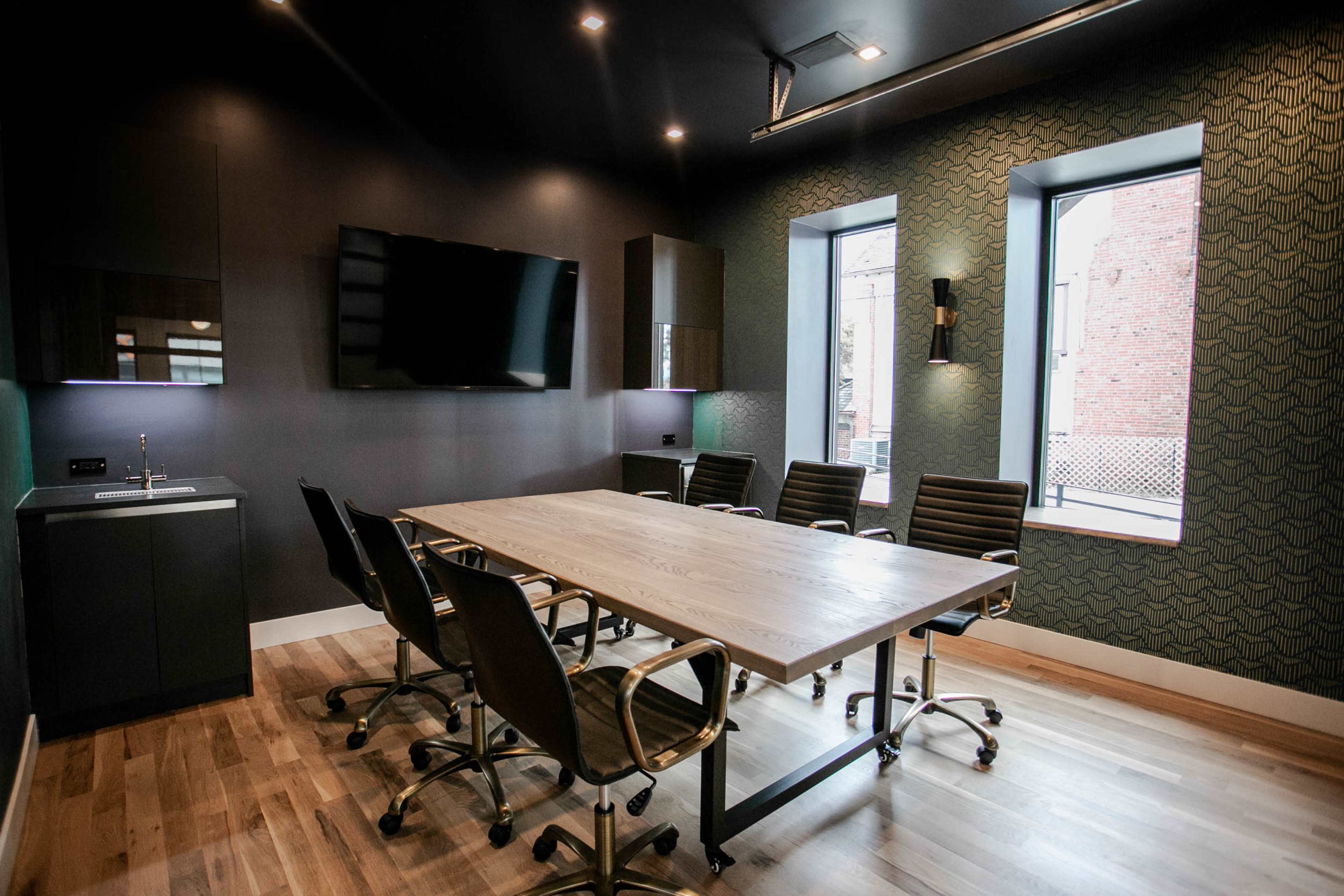 The image features a modern conference room with a large wooden table surrounded by black leather chairs, a wall-mounted television, and two windows.