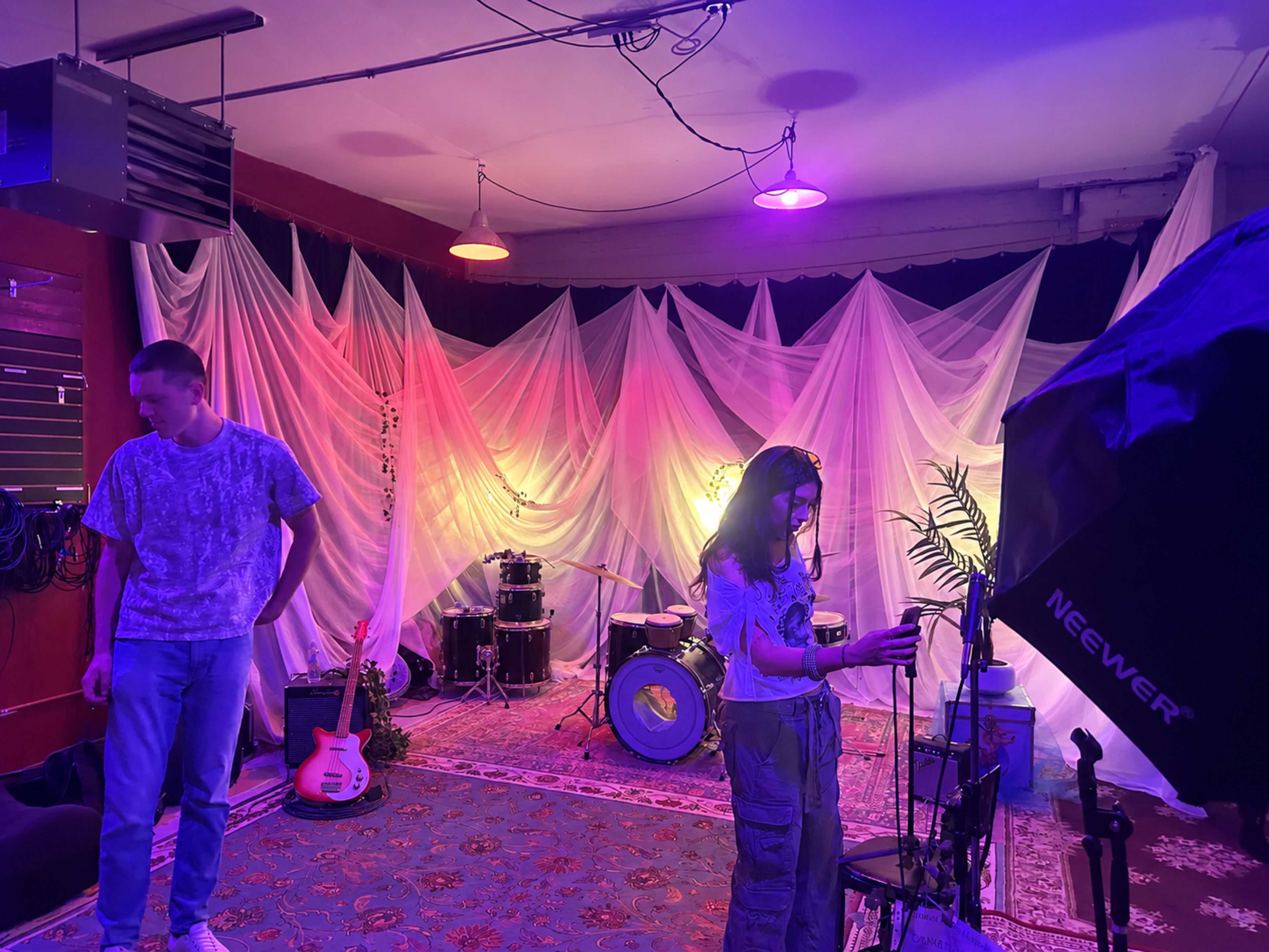 A young man stands beside a drum set and a young woman adjusts a camera in a studio with colorful lights and draped fabric in the background.