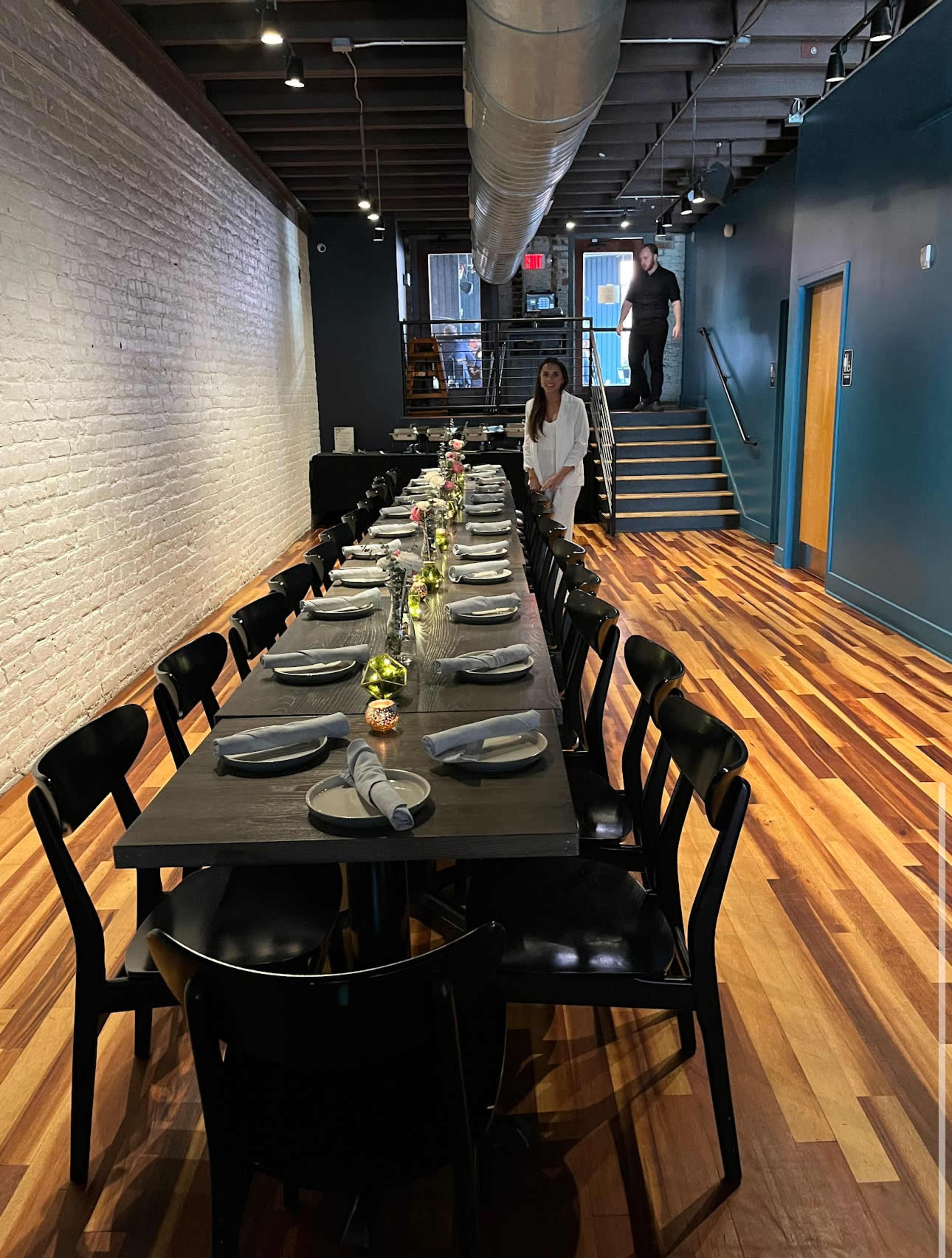 A long dining table is set with plates and utensils in a modern restaurant, while a woman prepares the table and a man walks up the stairs in the background.
