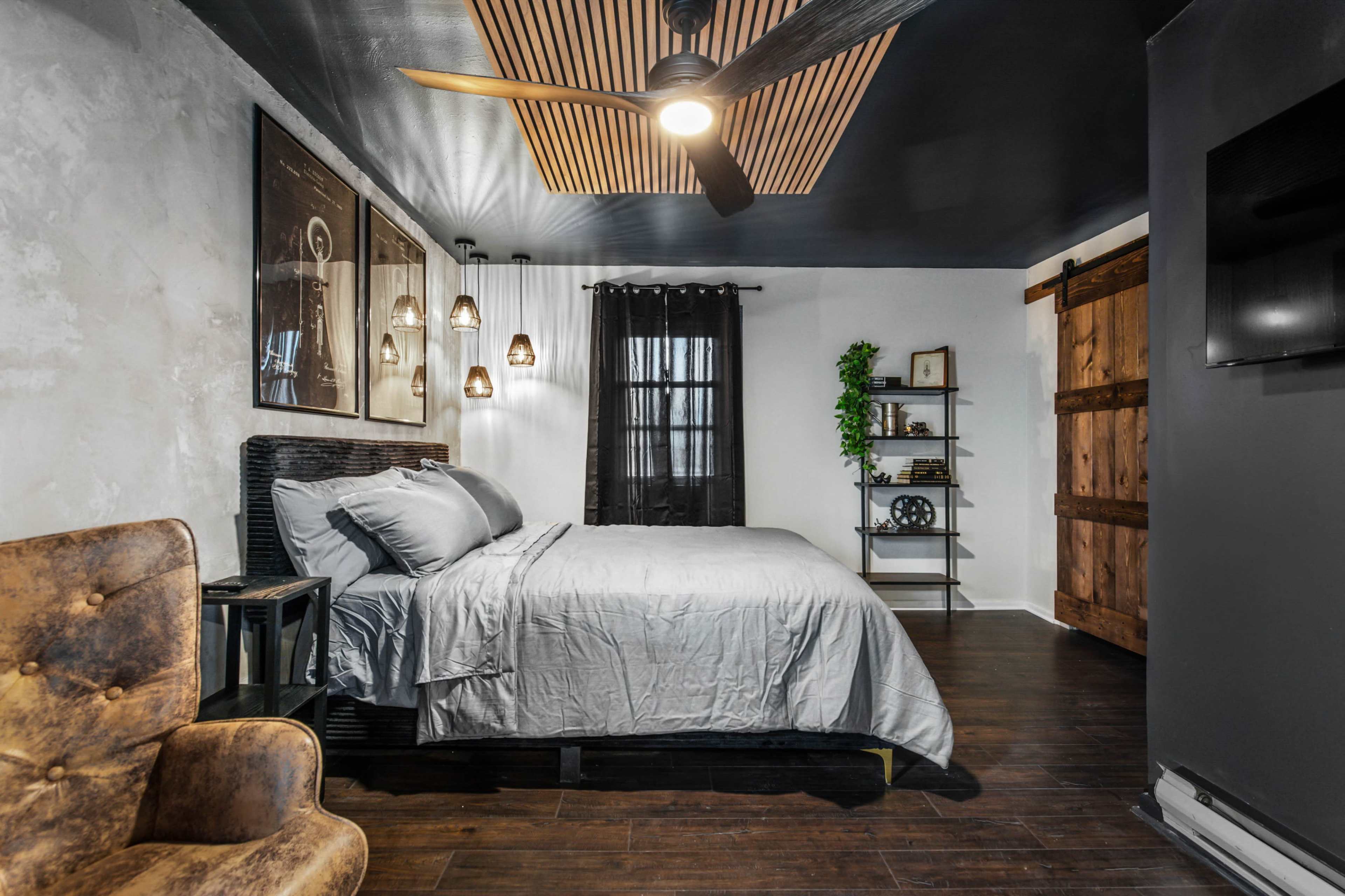 A modern bedroom with a gray bedspread, wooden accent walls, and a ceiling fan, featuring a cozy seating area and a small bookshelf.