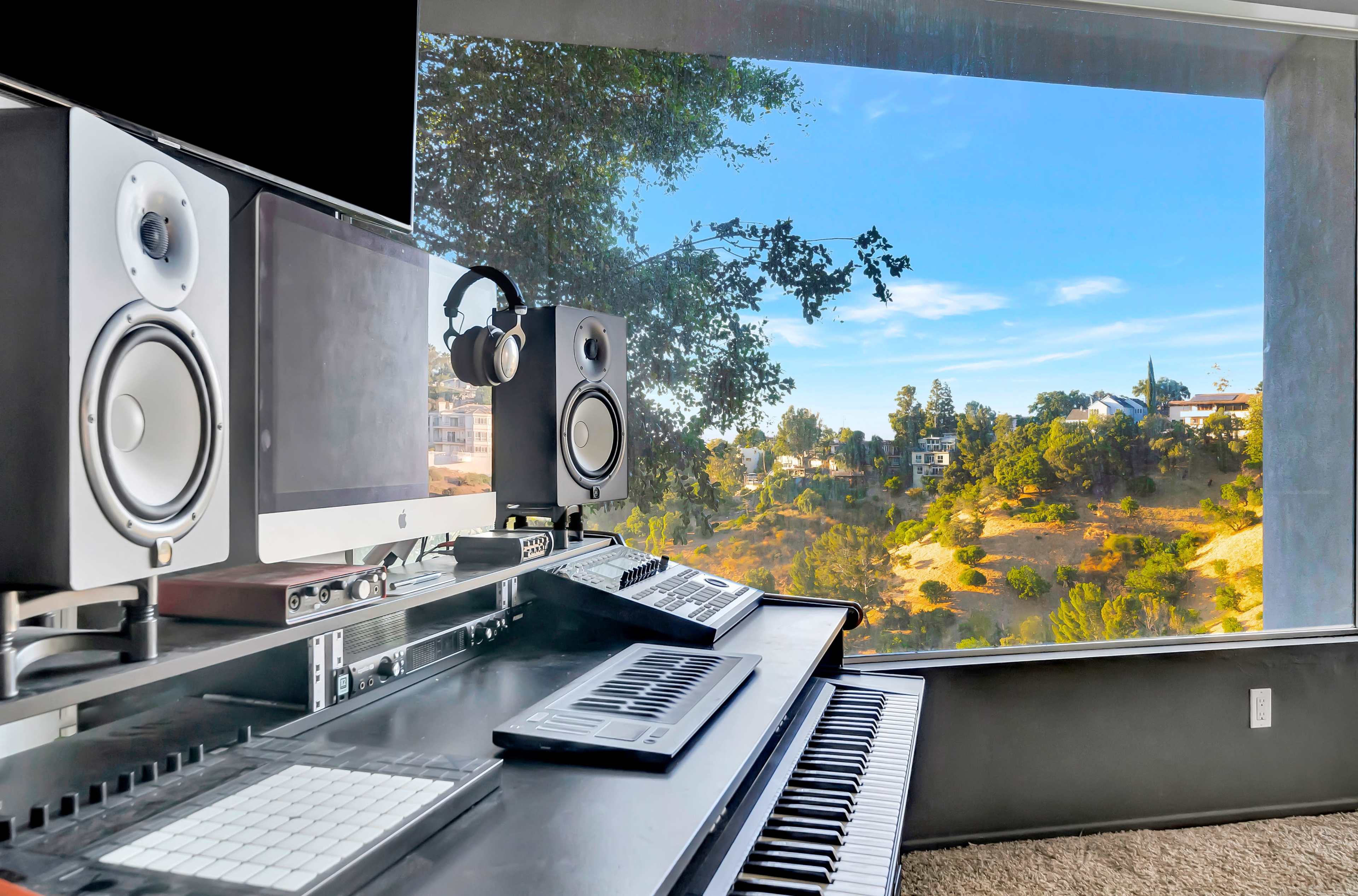 A music production workstation with speakers and a computer overlooks a landscaped hillside through large windows.