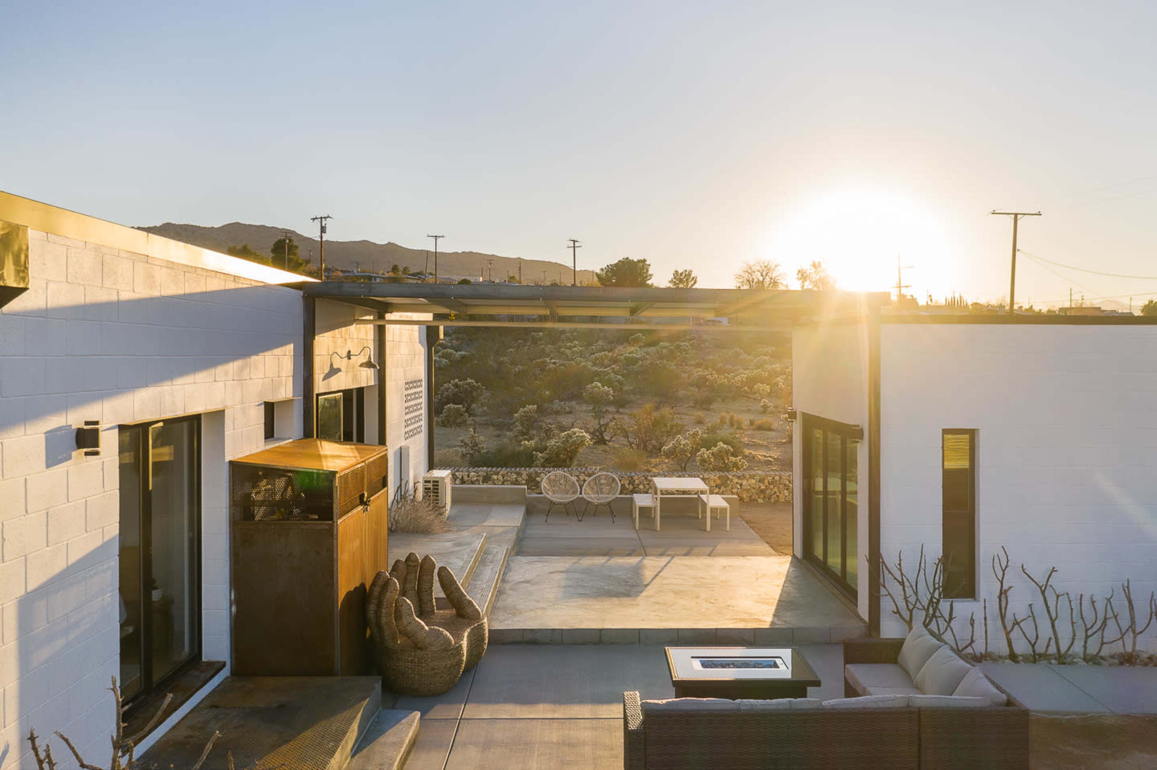 The image shows a modern architectural outdoor space with seating areas, a fire pit, and a sunset view over a desert landscape.