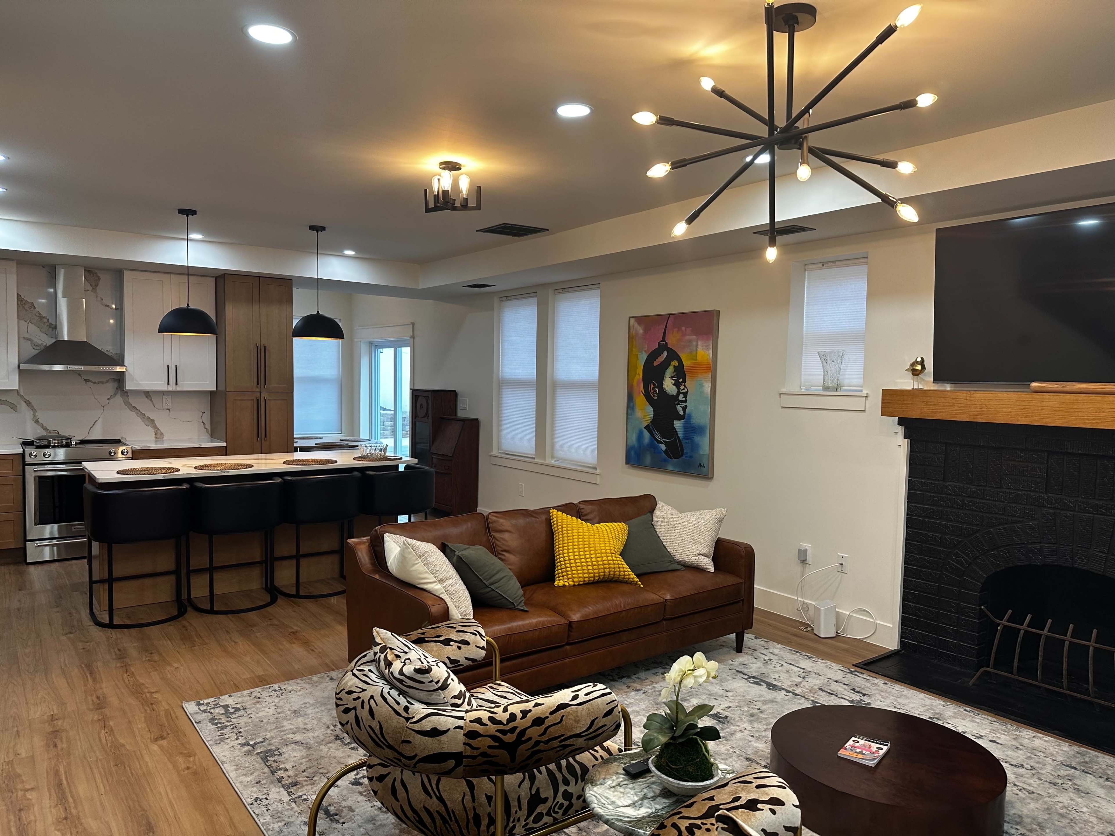 The image shows a modern living area featuring a brown leather couch, an abstract painting on the wall, and a round wooden coffee table, with a kitchen area visible in the background.