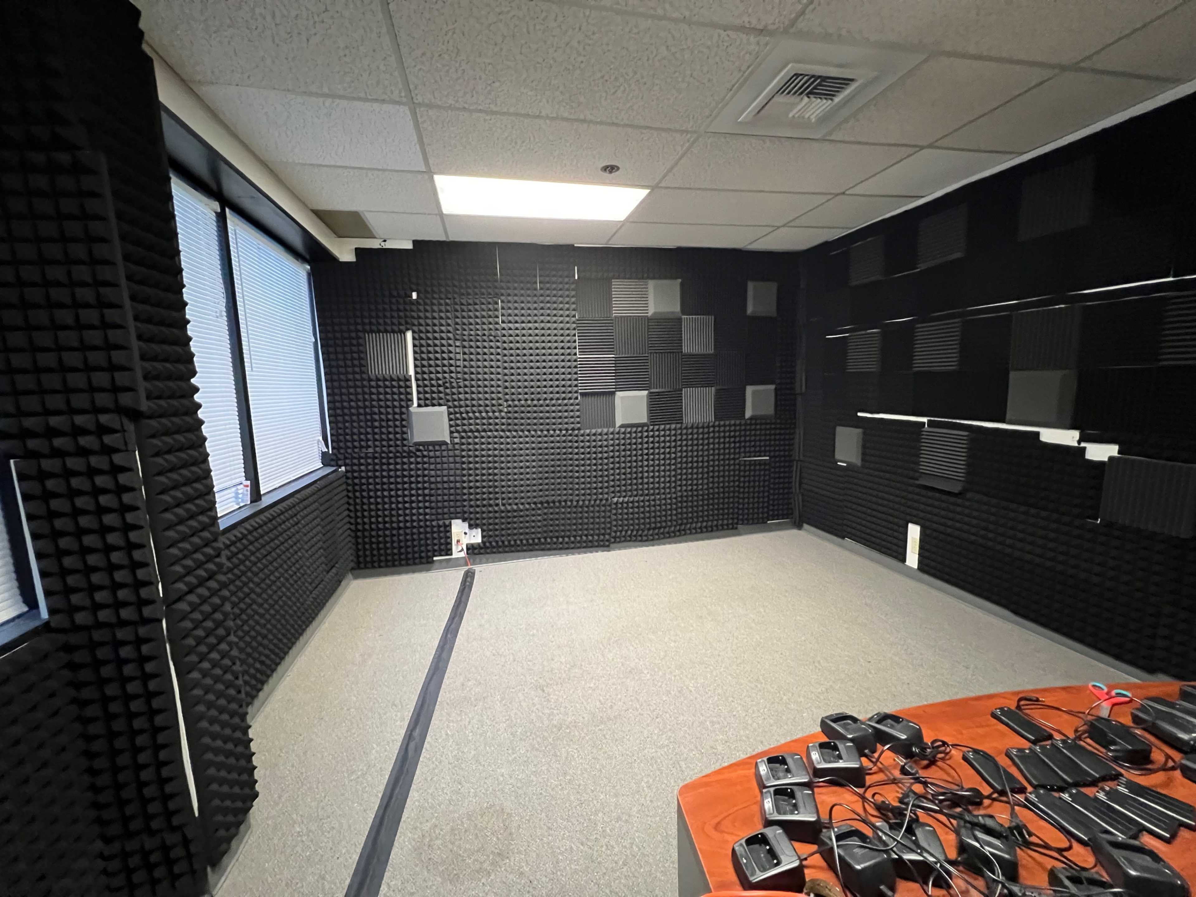 A soundproof room lined with black acoustic panels, featuring a gray carpet and a wooden table with electronic devices on one side.