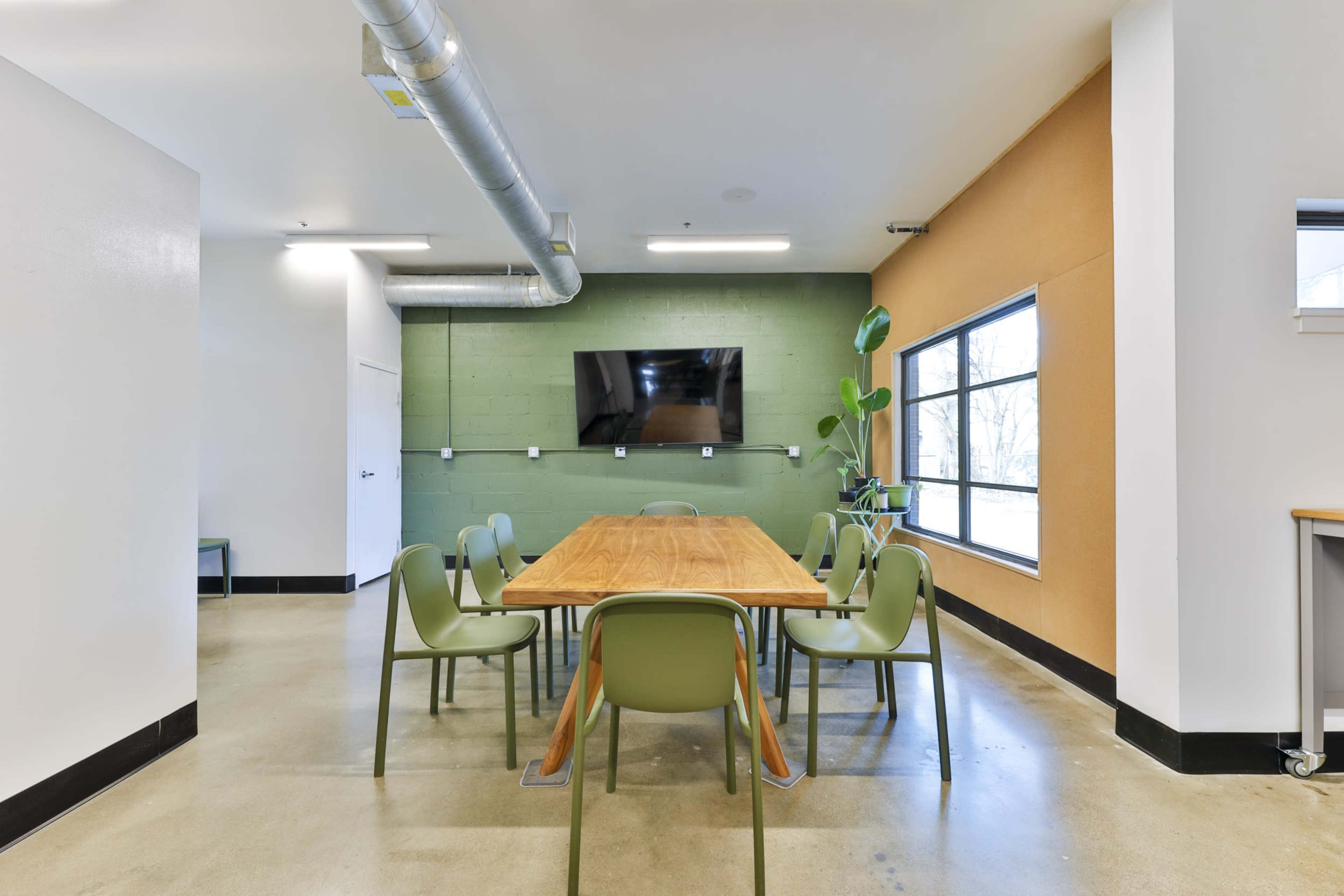 A long wooden table surrounded by green chairs is set in a meeting room with a large wall-mounted screen and windows.
