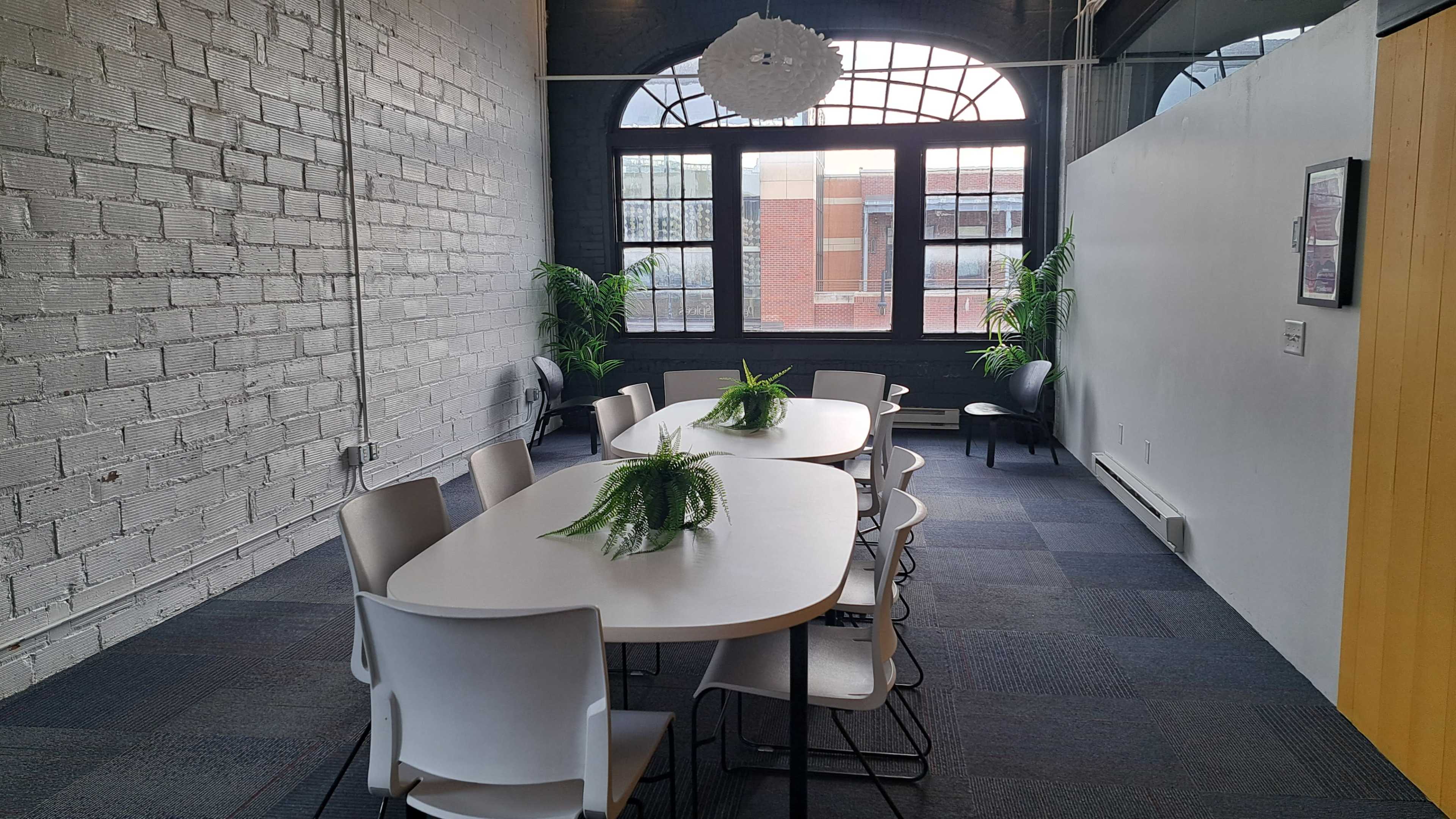 The image shows a conference room with a long, white table surrounded by chairs, accented by potted plants and large windows.