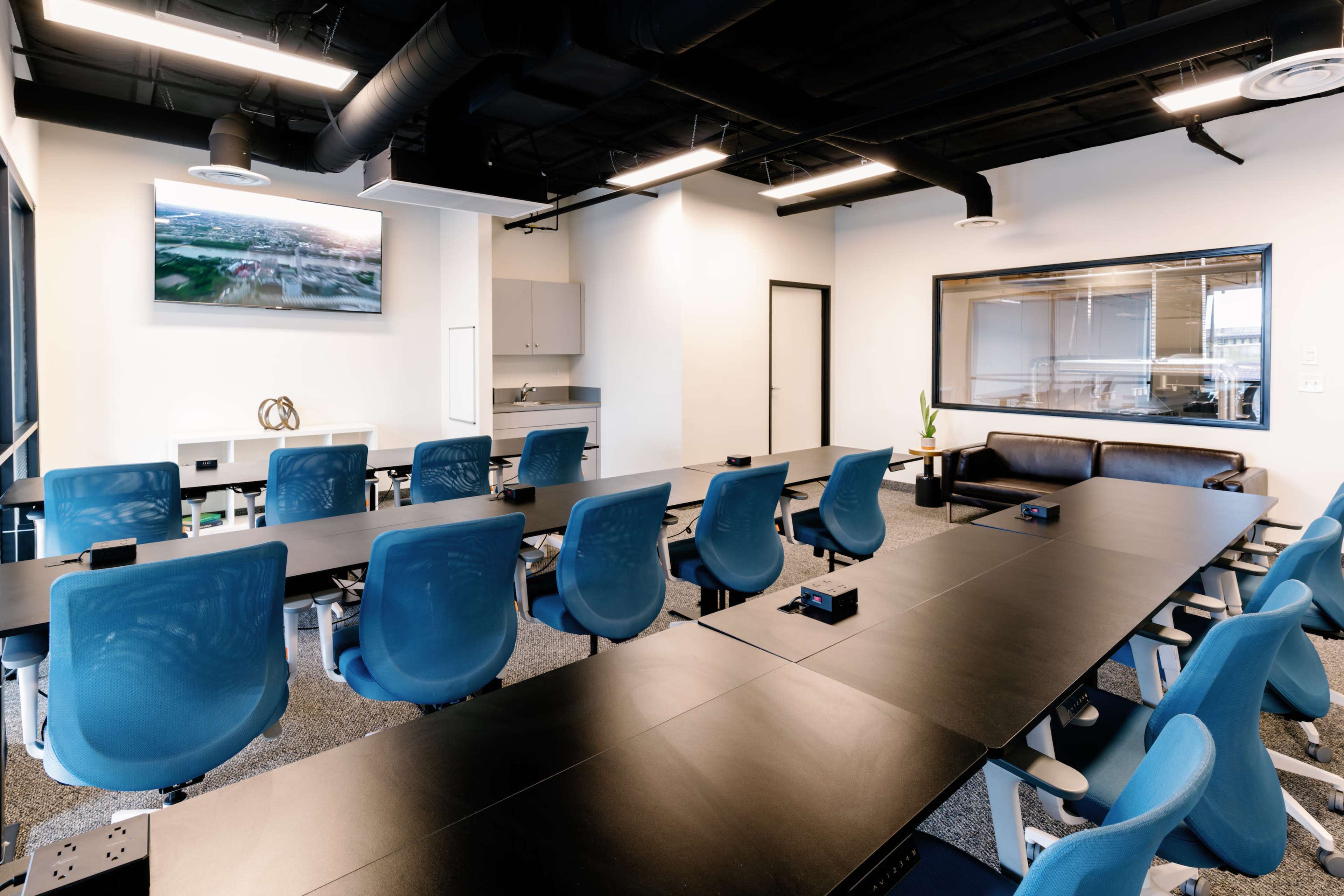 A modern conference room features a long table with blue chairs, a wall-mounted screen, and a kitchenette in the background.