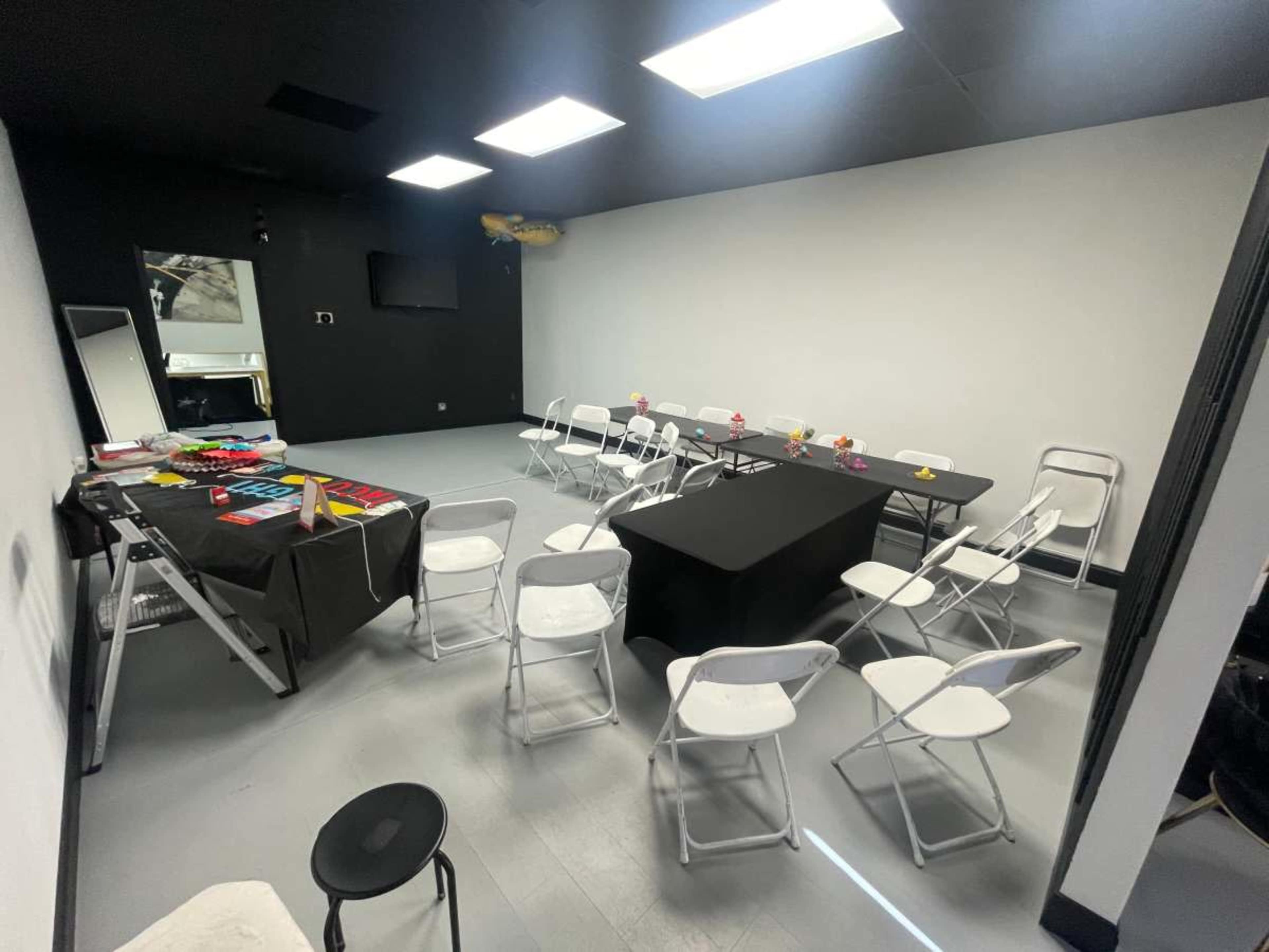 The image shows a neatly arranged meeting room with a long black table, several white folding chairs, and a table covered with colorful materials, all set against a plain wall and bright overhead lighting.