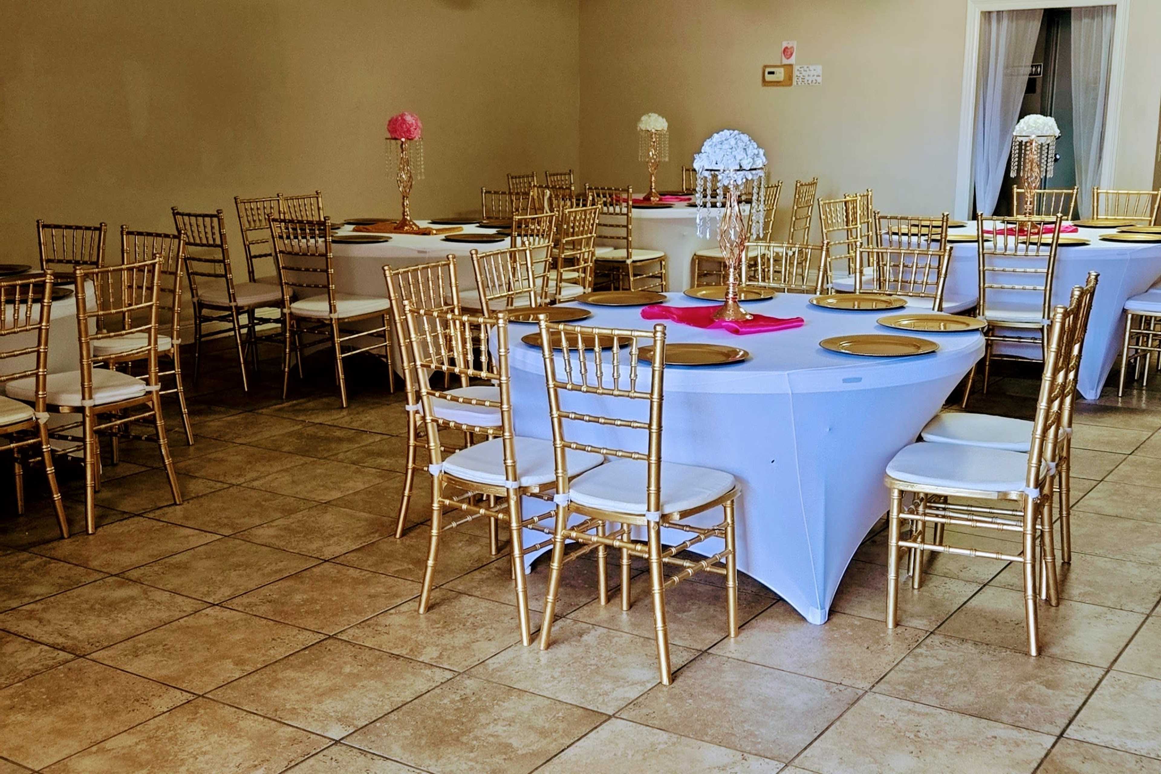 The image shows a banquet hall with multiple round and rectangular tables, each set with gold plates and adorned with decorative centerpieces, surrounded by gold-colored chairs.
