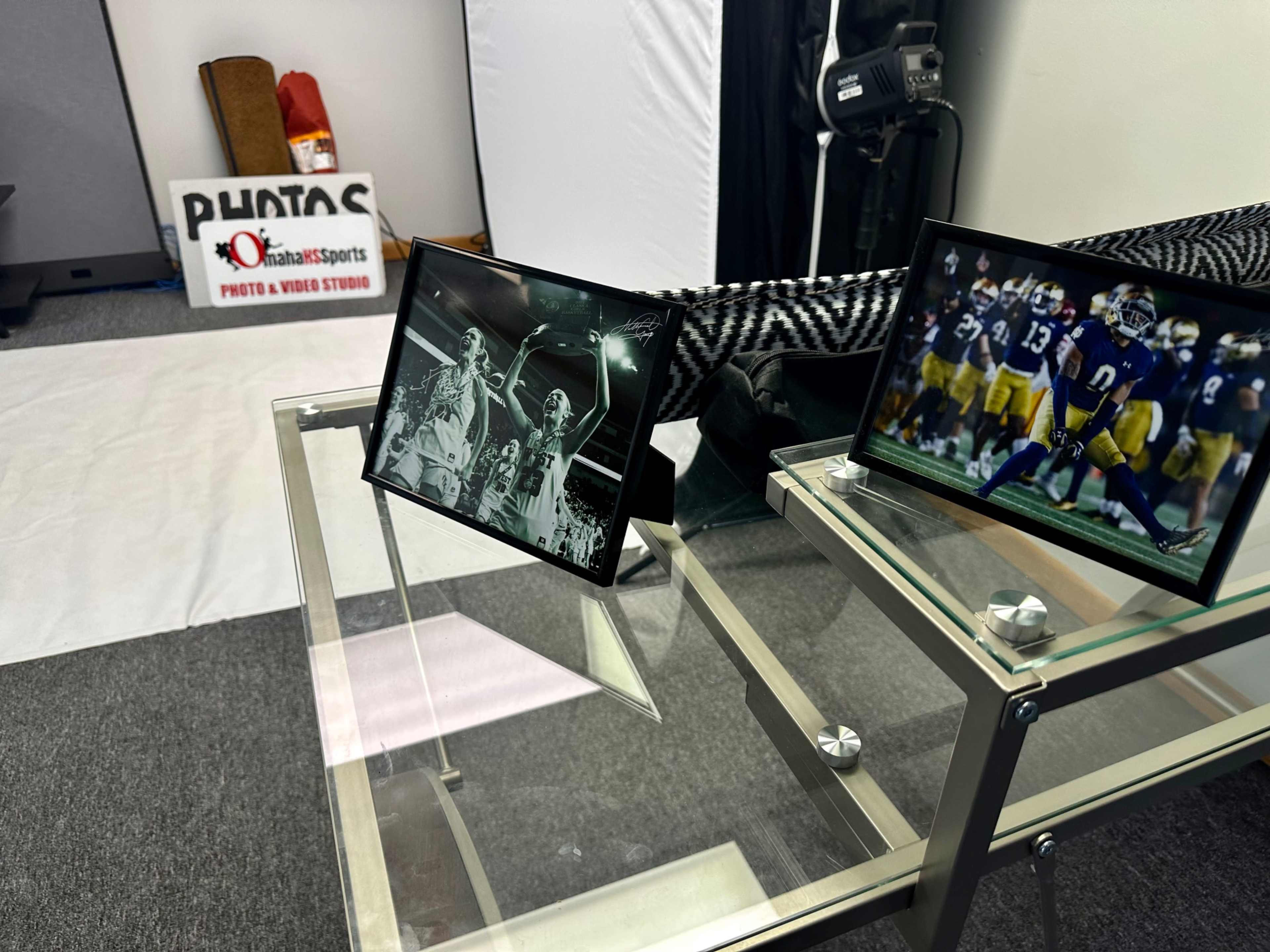 The image shows a glass table with two framed photographs, one depicting a sports celebration and the other featuring a football player, set in a photo and video studio.