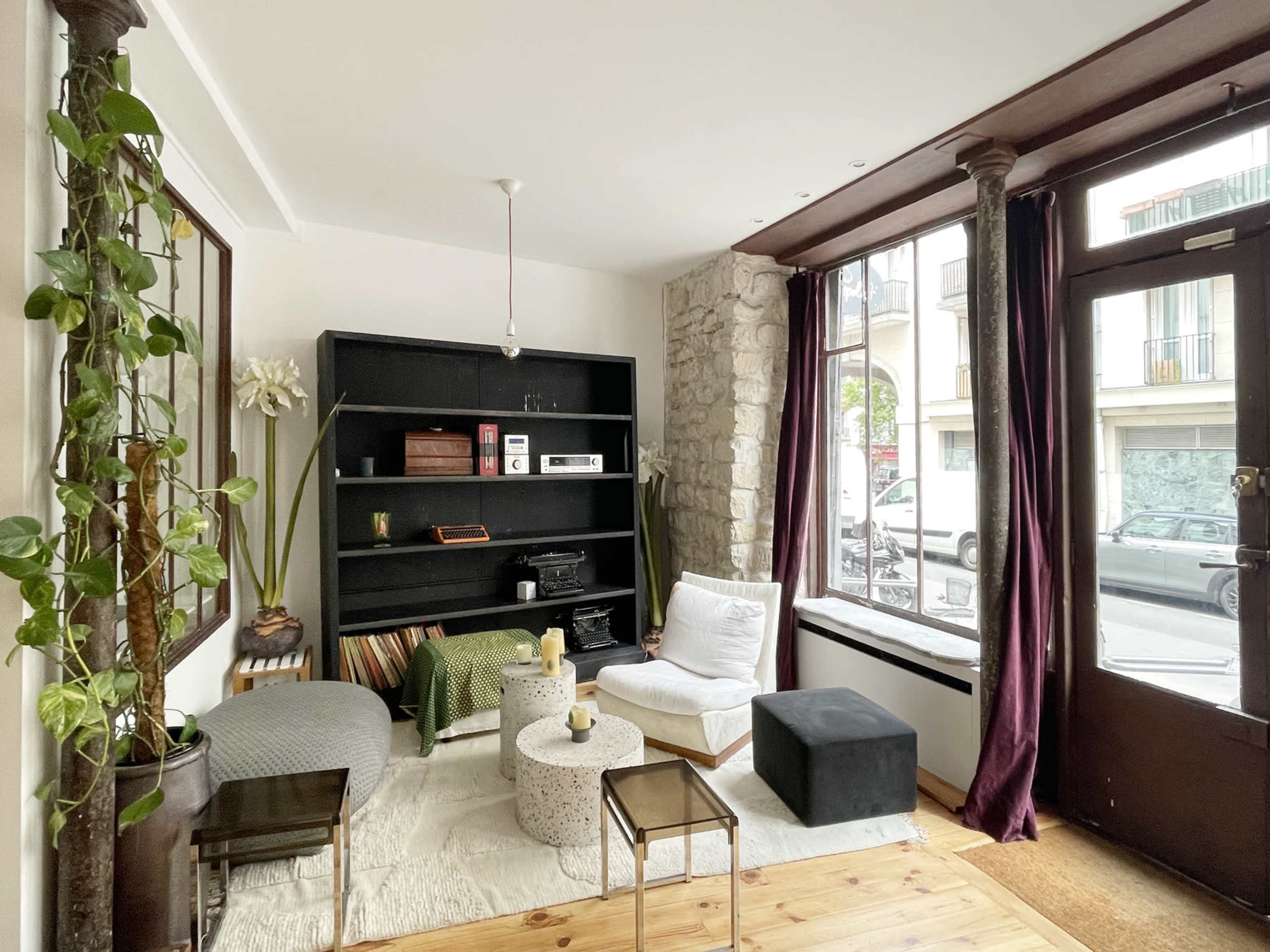 A cozy living room with a black bookshelf, minimalist furniture, a coffee table, and large windows allowing natural light to enter.