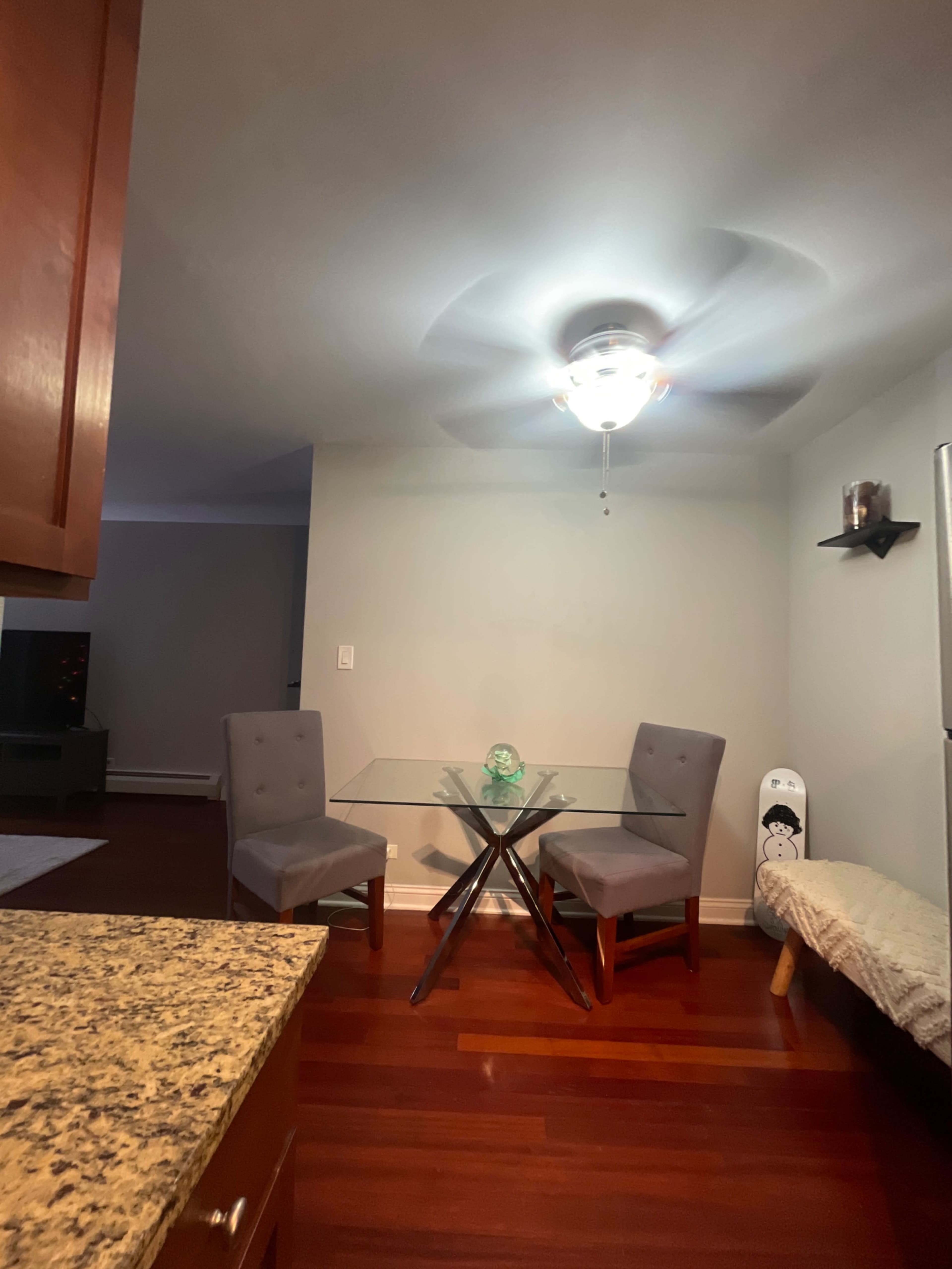 A modern dining area features a glass table with two gray chairs and a ceiling fan, surrounded by hardwood flooring.