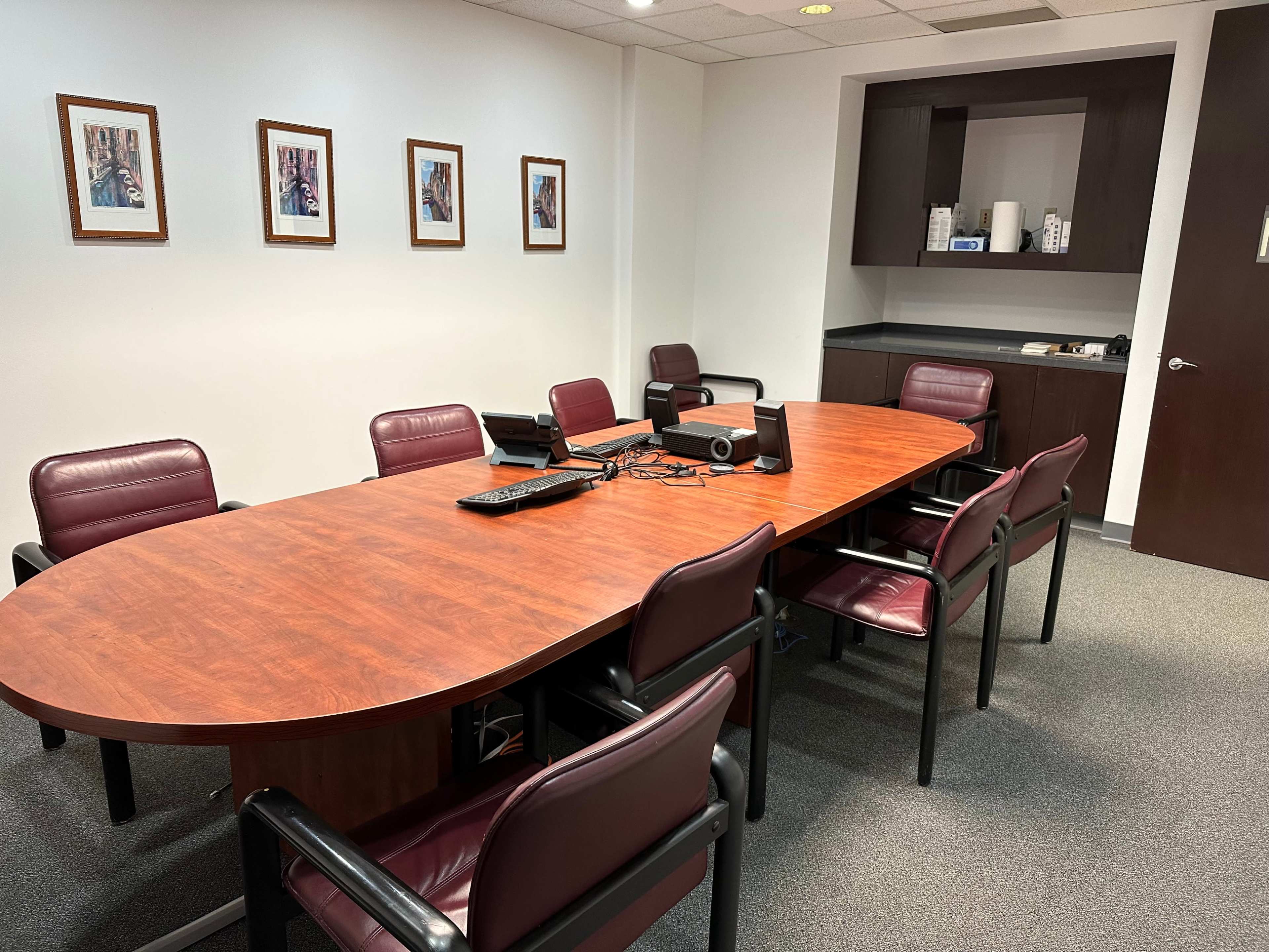 The image shows a conference room with an oval wooden table surrounded by six maroon chairs, framed pictures on the walls, and a black cabinet in the corner.