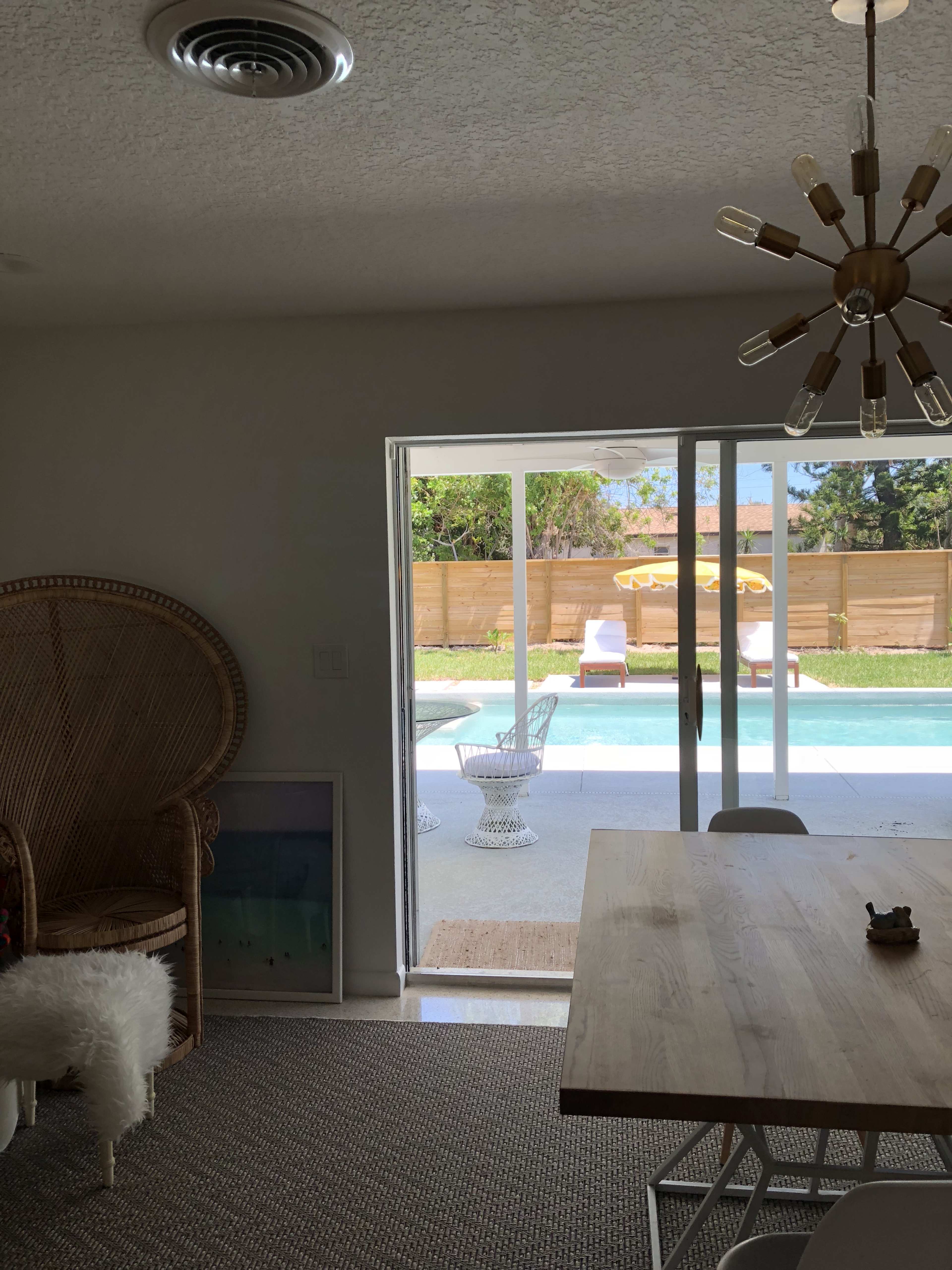 A spacious indoor area features a dining table and a large chair, with sliding glass doors opening to a patio and pool area.