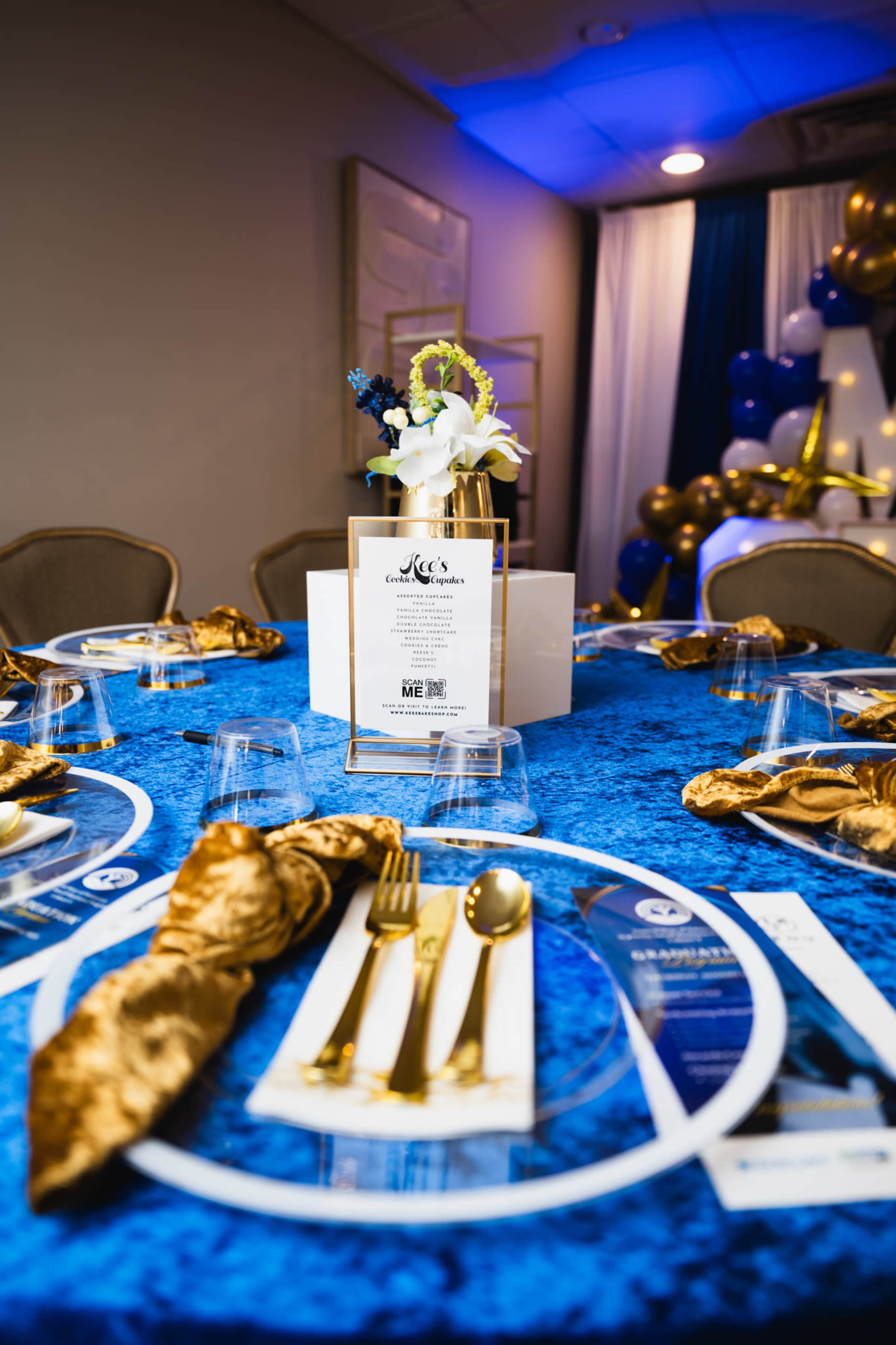 The image shows a beautifully arranged dining table with a blue tablecloth, elegant gold napkins, a floral centerpiece, and a menu displayed prominently in the center.
