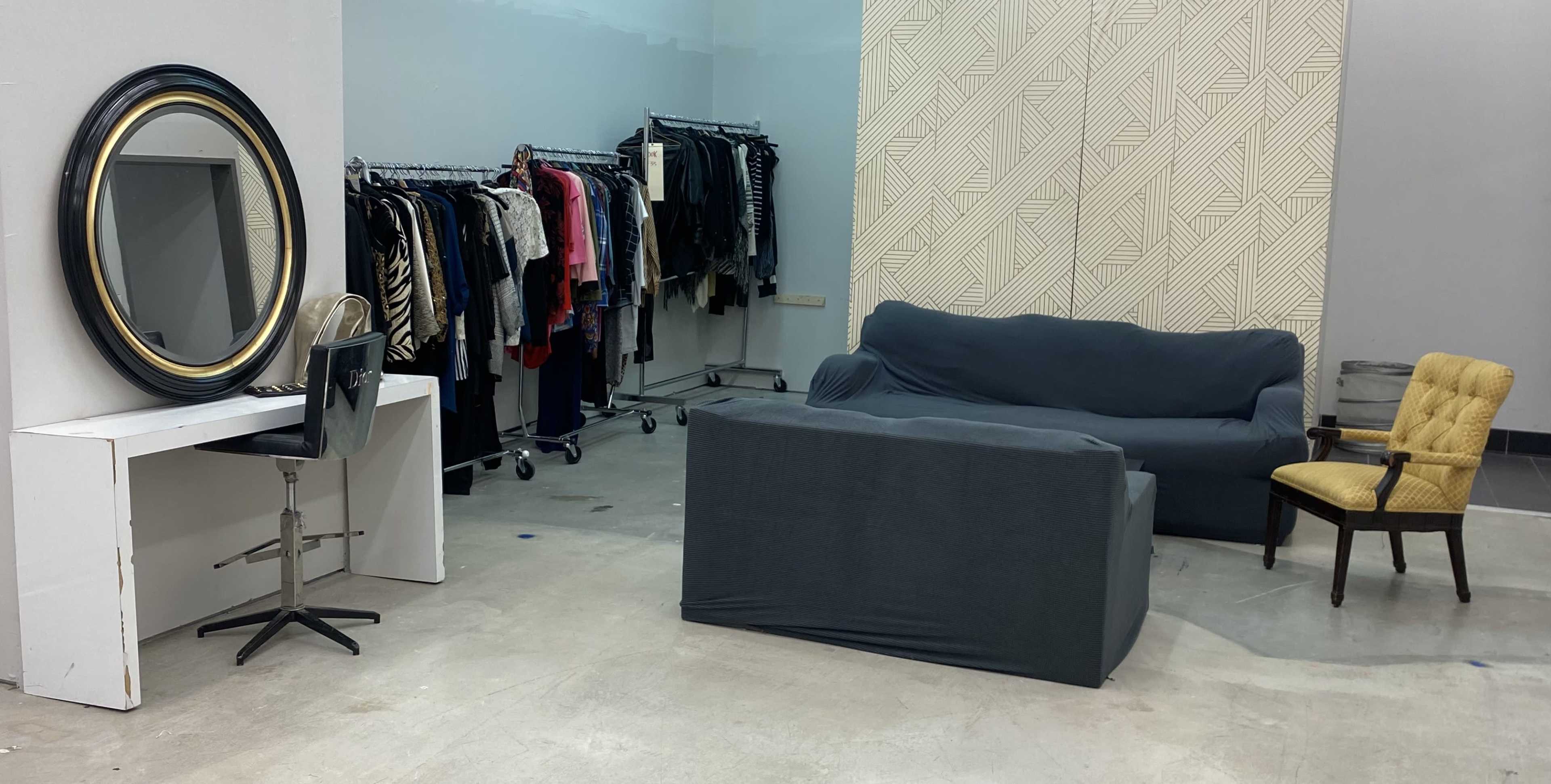 The image shows a dressing area with a black sofa, a yellow chair, a mirror on a white table, and a rack of clothing in the background.