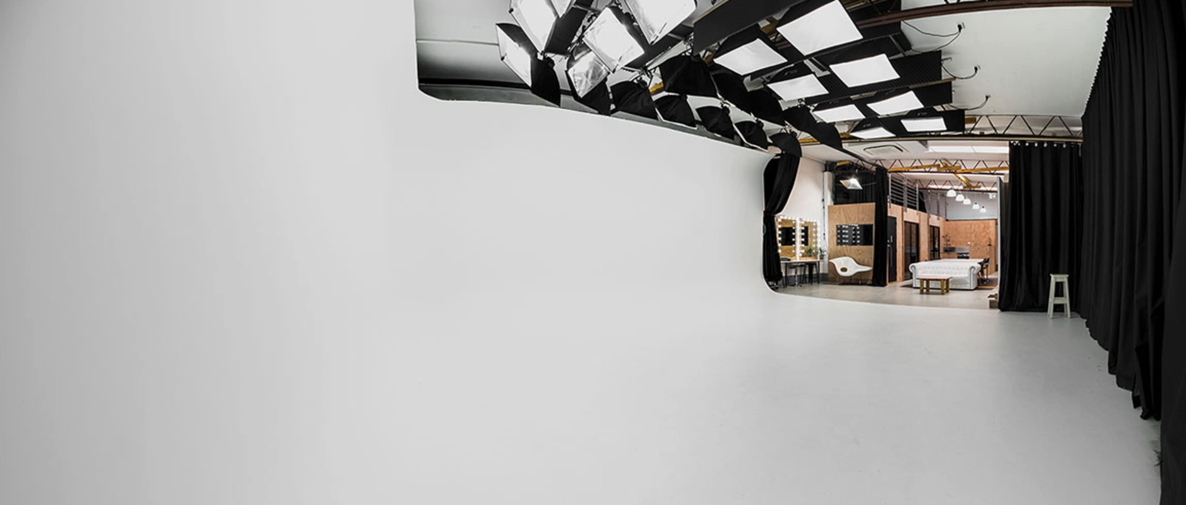 A spacious studio with a seamless white backdrop, overhead lighting fixtures, and a partitioned area featuring furniture and a mirror setup.