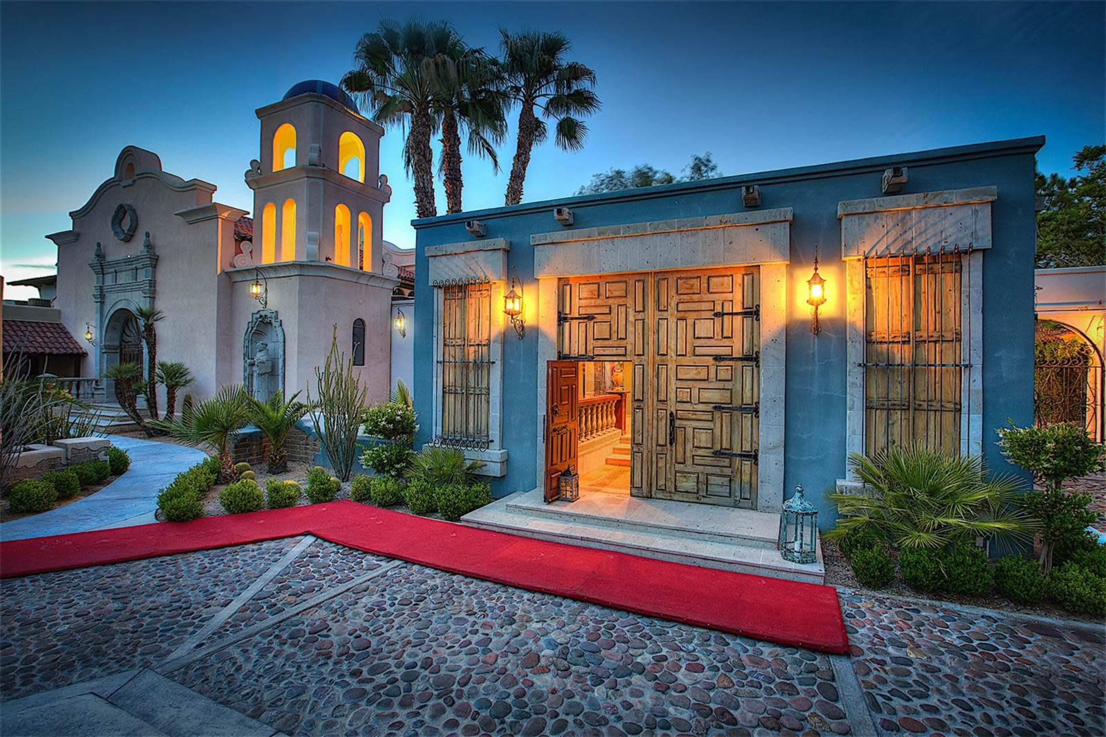 A beautifully lit entrance to a building with ornate doors and a red carpet, surrounded by tropical plants and stone pathways.