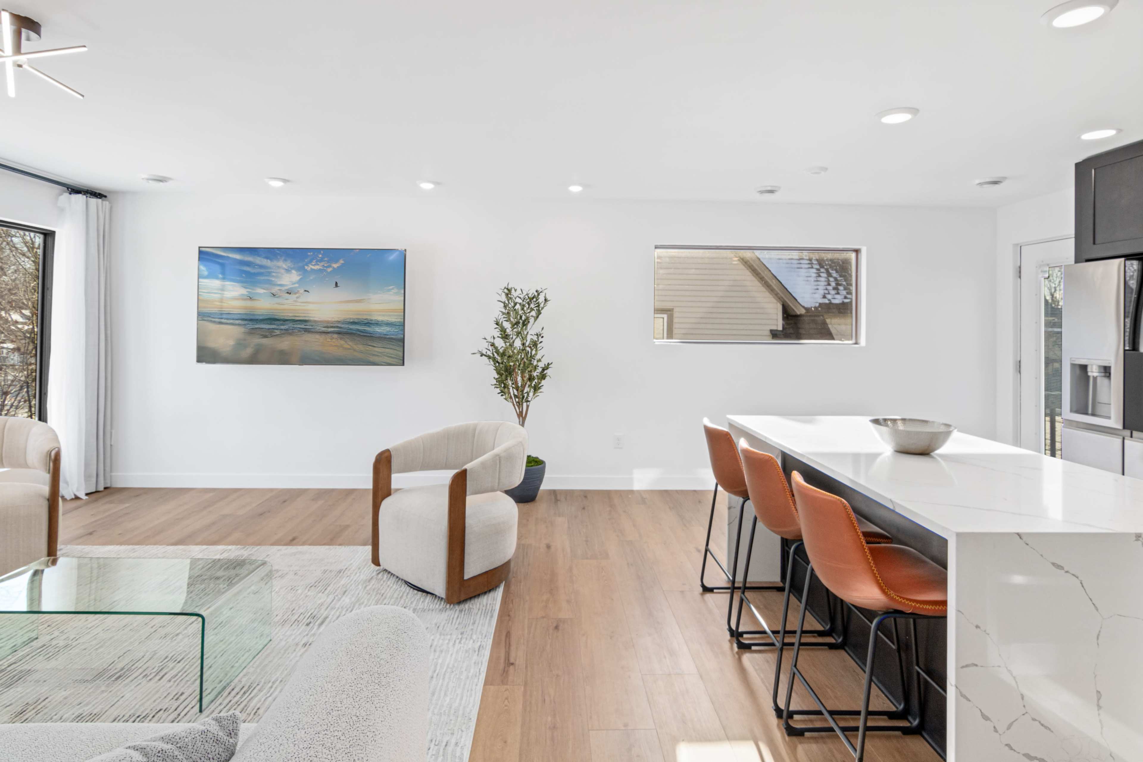 The image shows a modern living room with a dining area, featuring a large wall-mounted artwork, a glass coffee table, and four barstools at a sleek kitchen island.