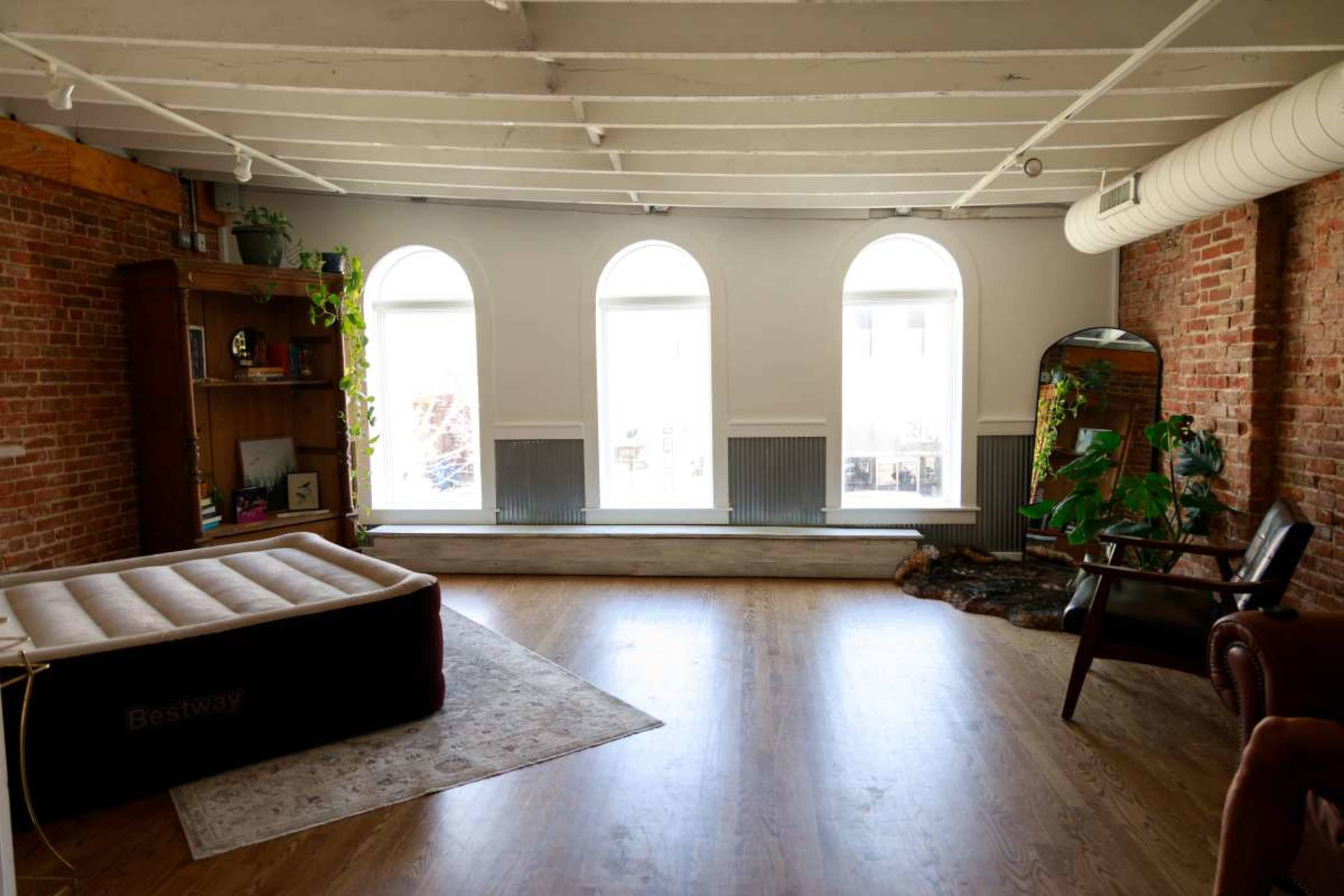 The image shows an open, spacious room with exposed brick walls, a large window with three arches, a brown air mattress, a wooden bookshelf, and indoor plants.