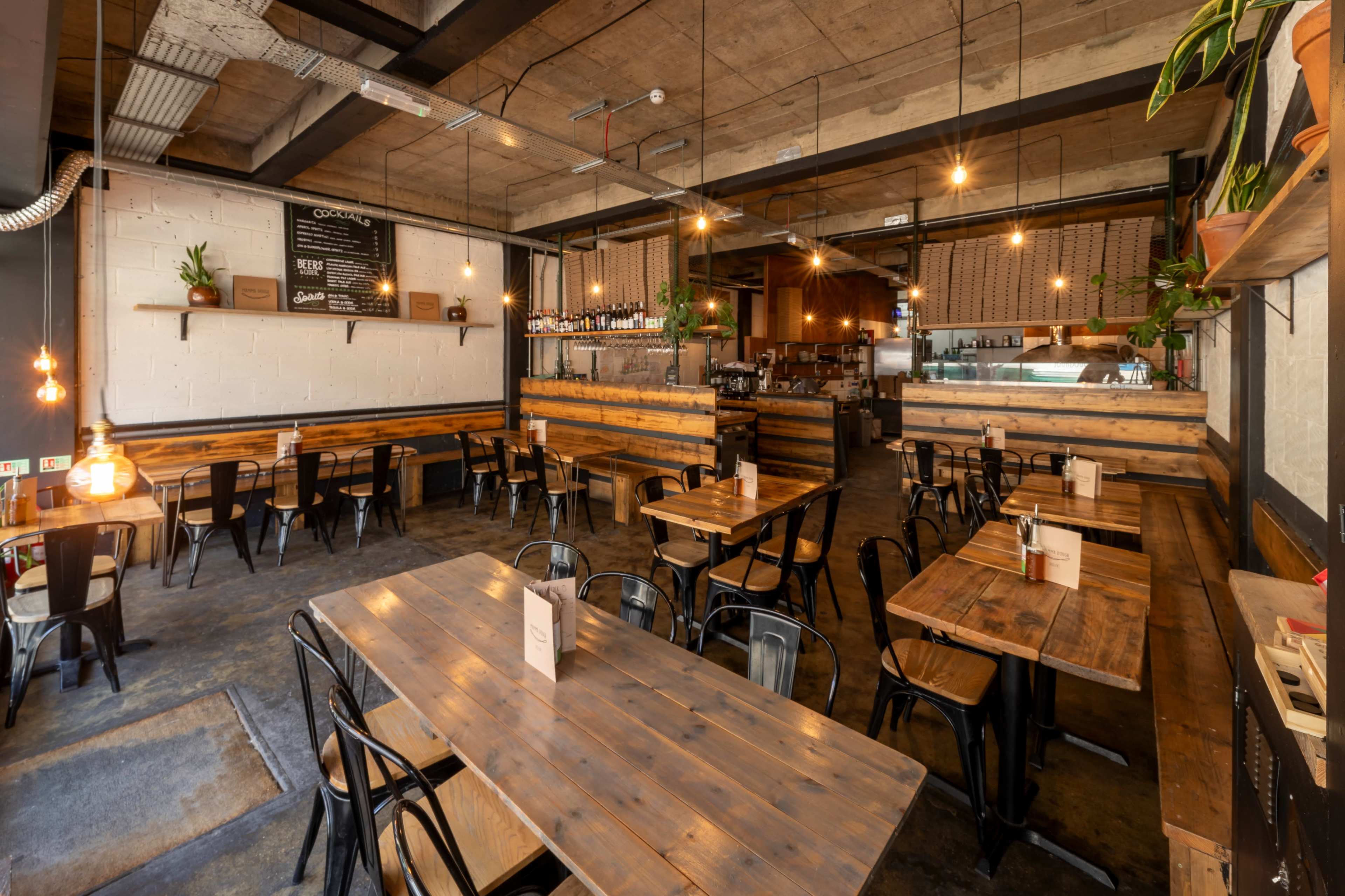The image shows a modern restaurant interior with wooden tables and metal chairs arranged around a spacious layout, featuring dim lighting and potted plants.