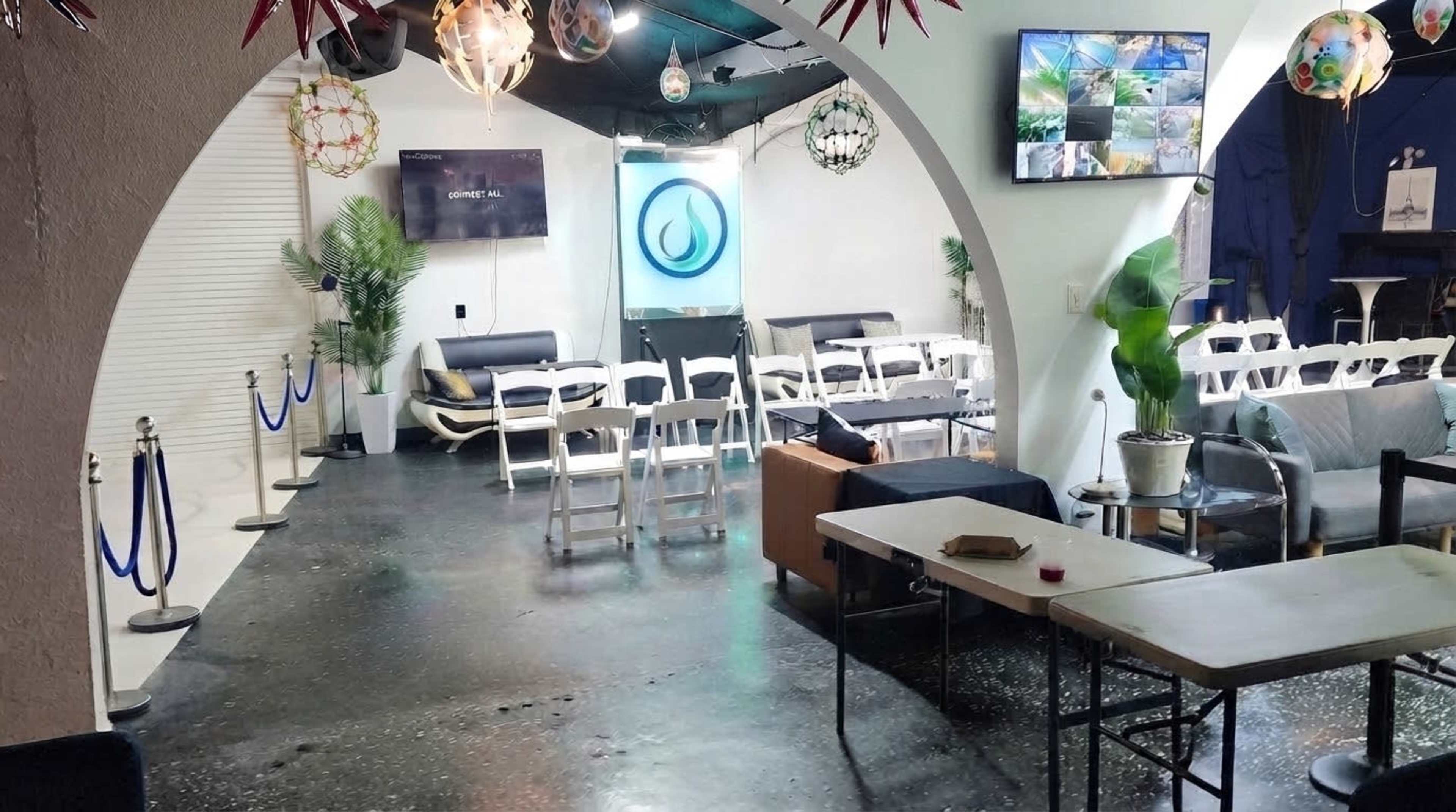 The image shows a spacious indoor area with seating arranged in rows, decorative lighting fixtures, and a large wall-mounted screen displaying various visuals.