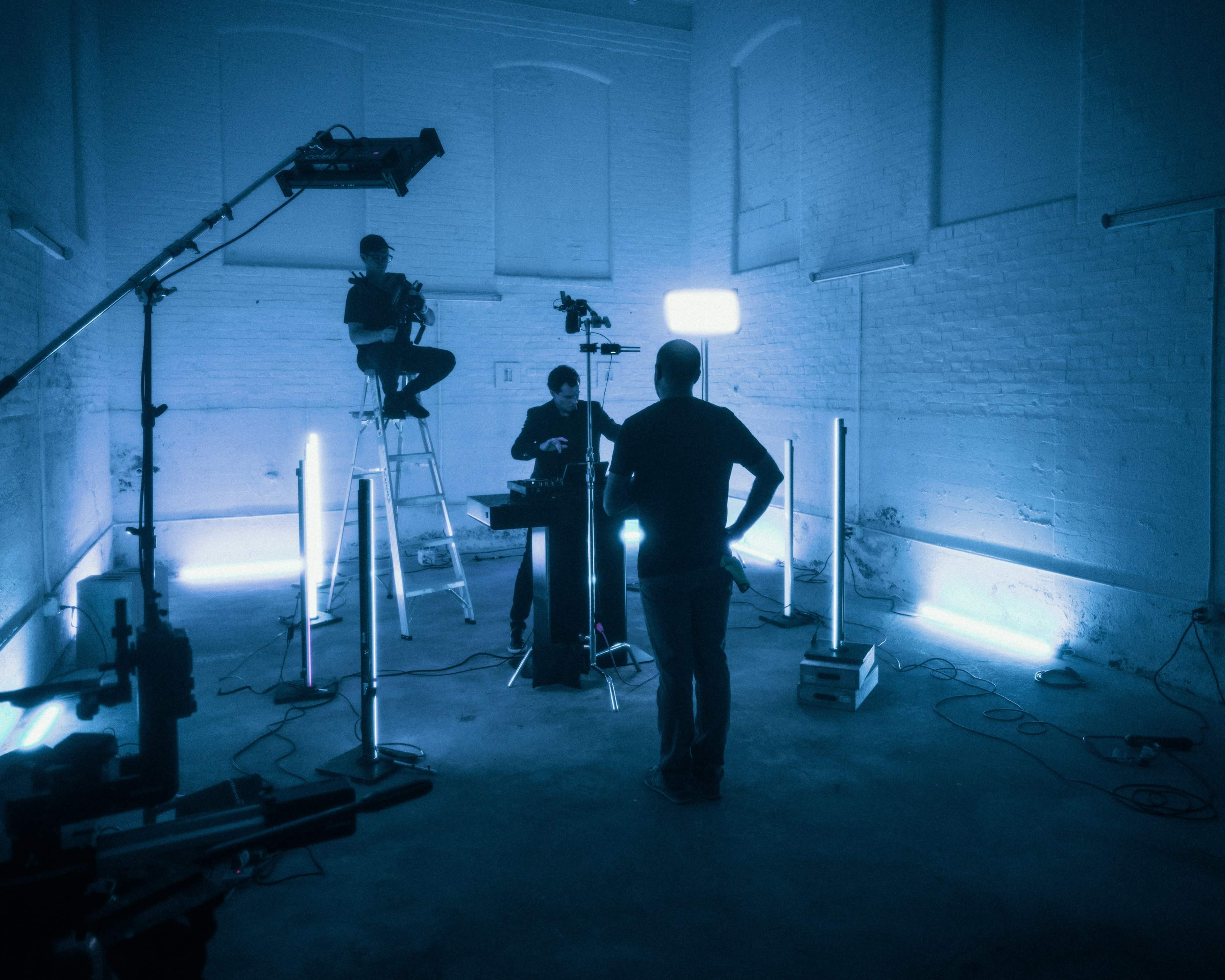 A film crew sets up equipment in a dimly lit studio featuring blue lighting.