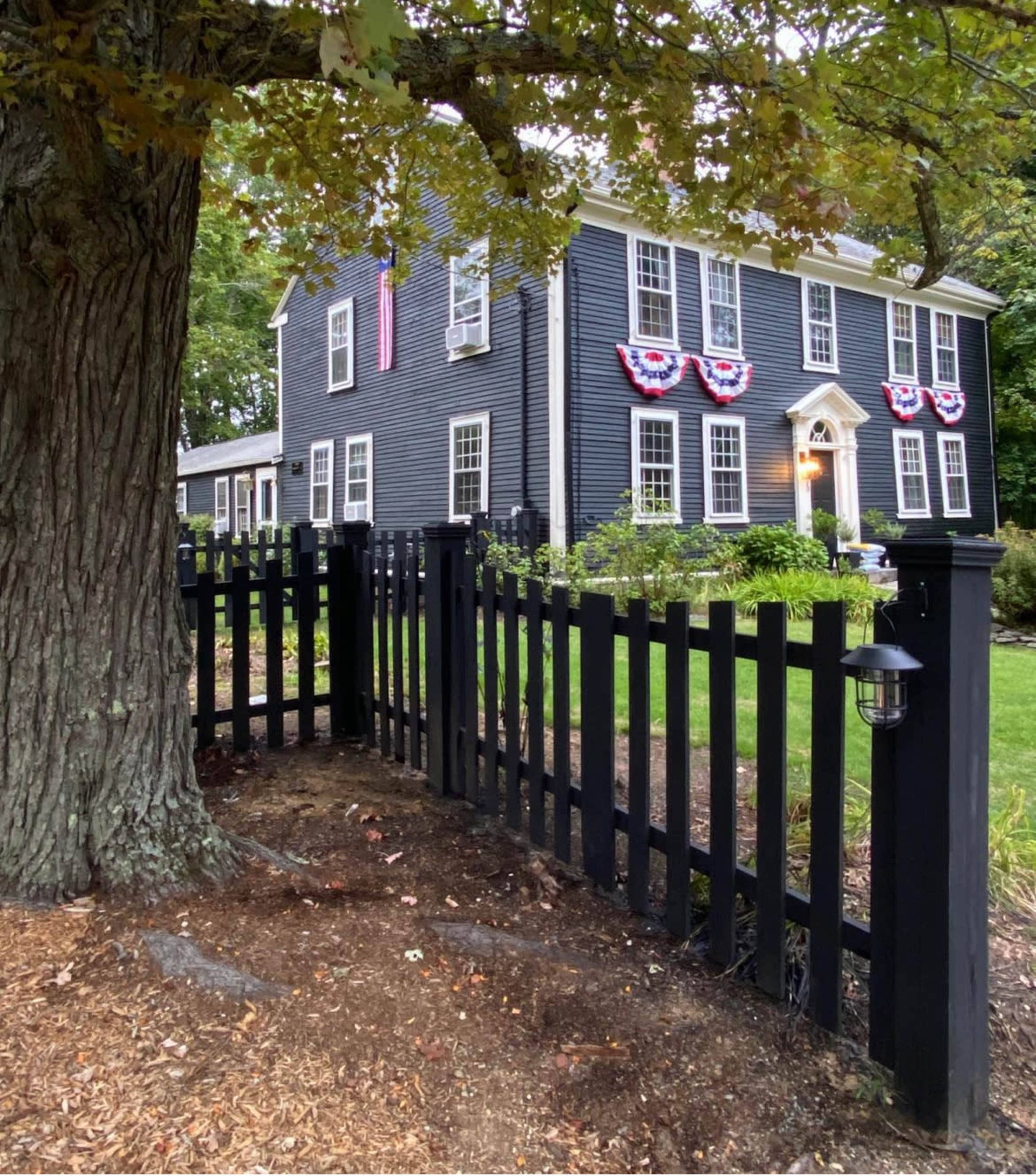 A black picket fence surrounds a two-story dark blue house that is adorned with red, white, and blue bunting.