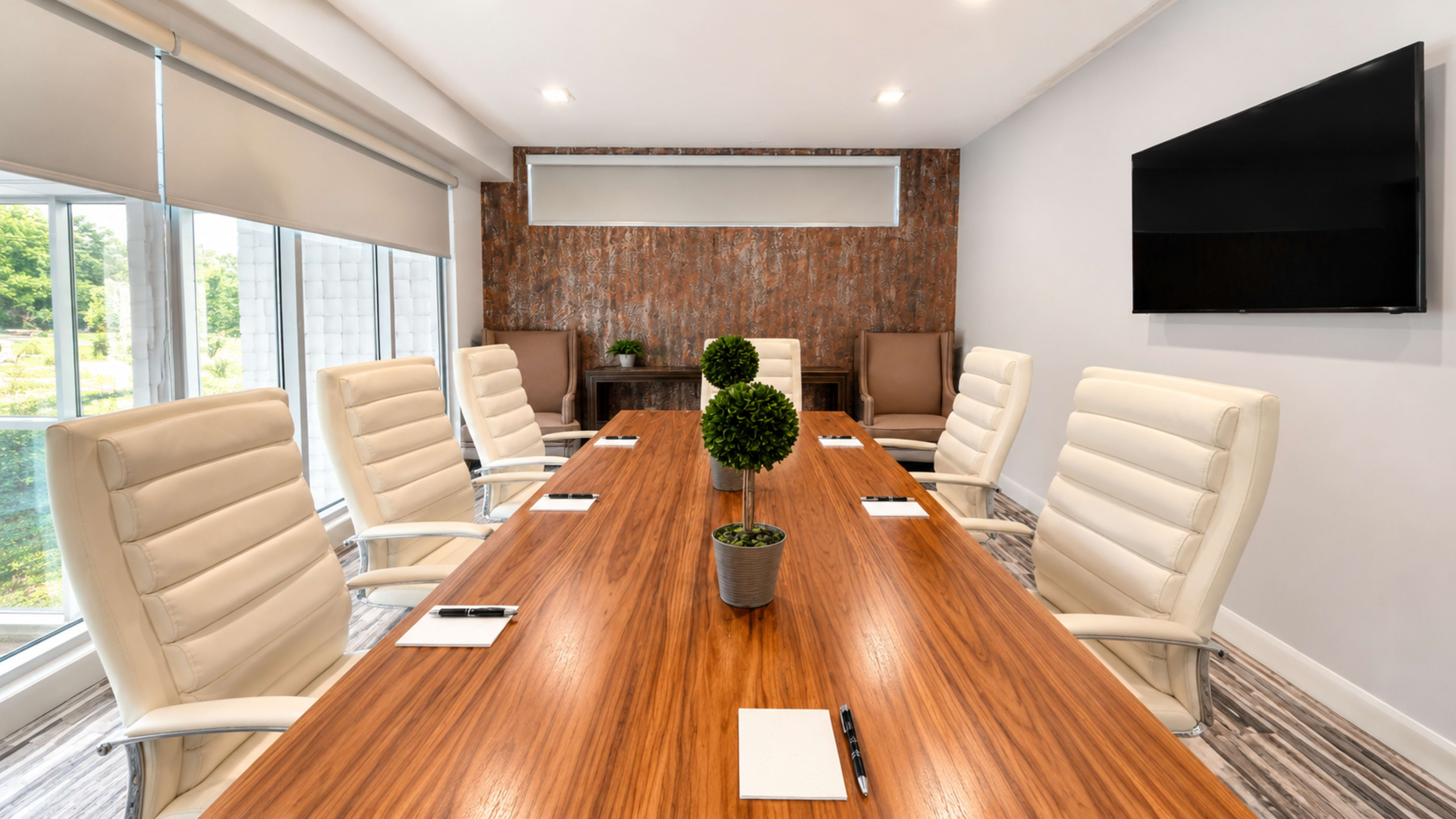 A large conference room features a wooden table surrounded by white leather chairs, with a wall of textured wood and a mounted television in the background.