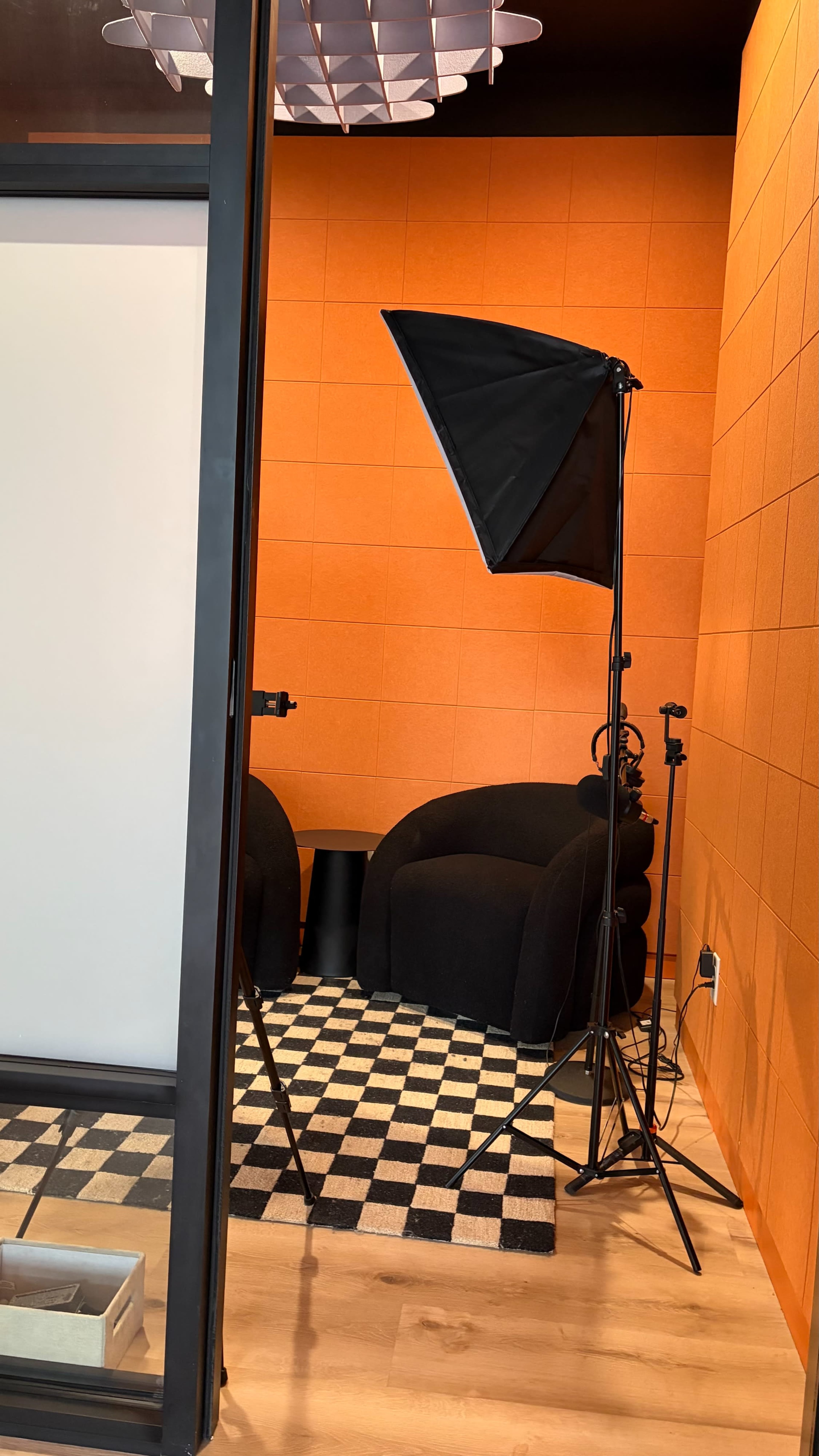 The image shows a small studio space with an orange wall, a black chair, and photography equipment including a softbox and tripods on a checkered floor.