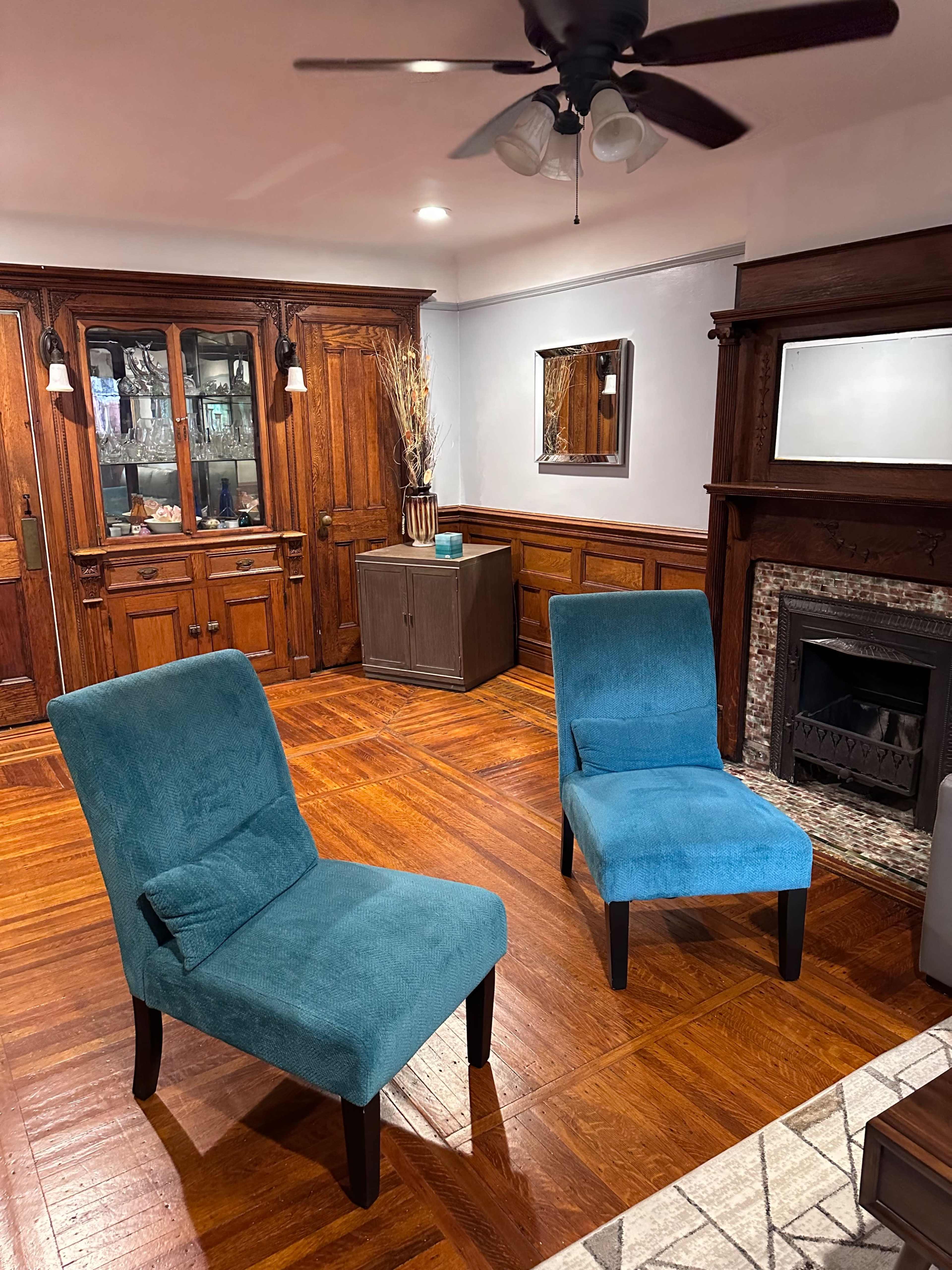The image shows two blue chairs positioned in a room with wooden paneling, a fireplace, and a glass-front cabinet.