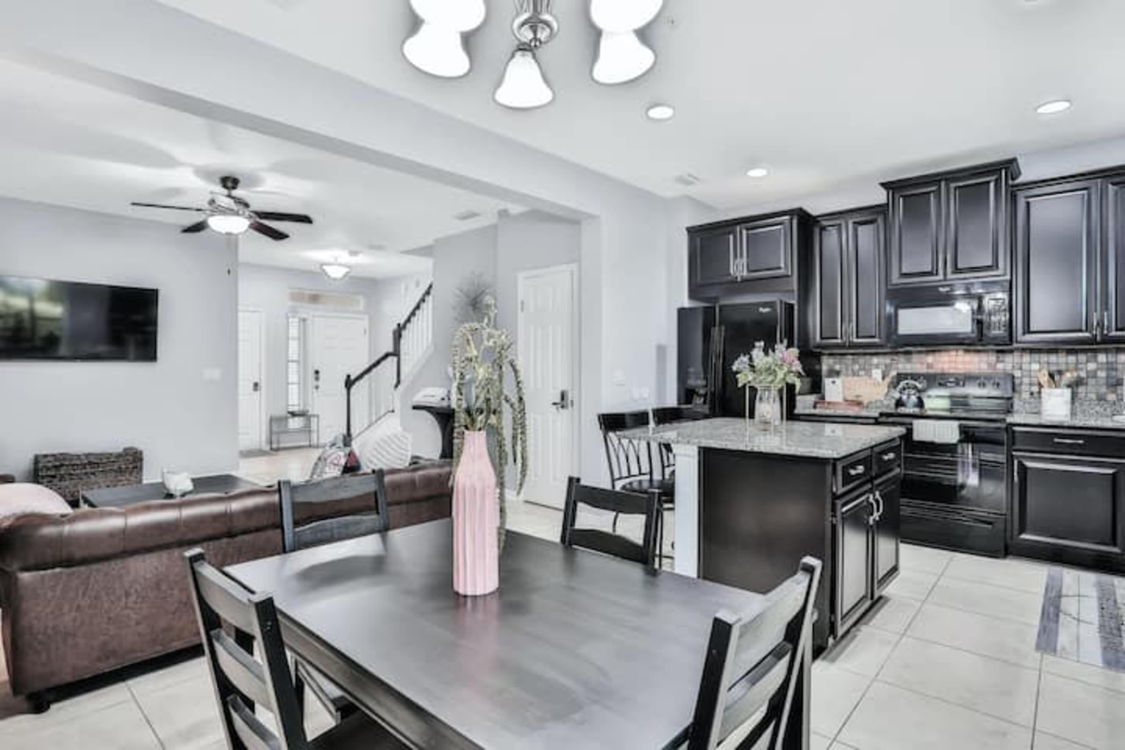 A modern open-concept kitchen and dining area features dark cabinetry, a granite countertop, and a large dining table with a vase of flowers.