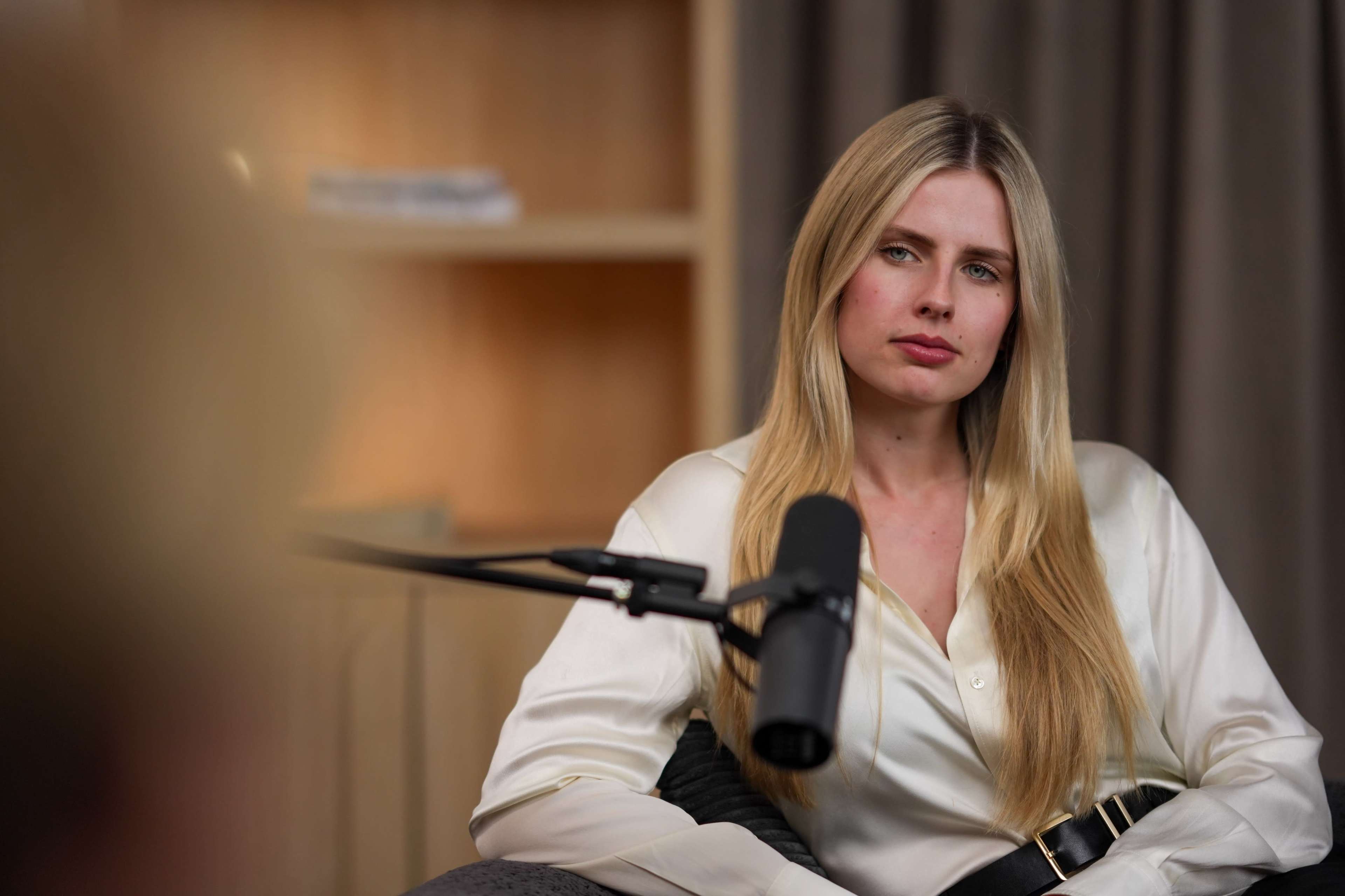 A woman with long blonde hair sits in a chair, looking intently at a person off-screen, with a microphone positioned nearby.