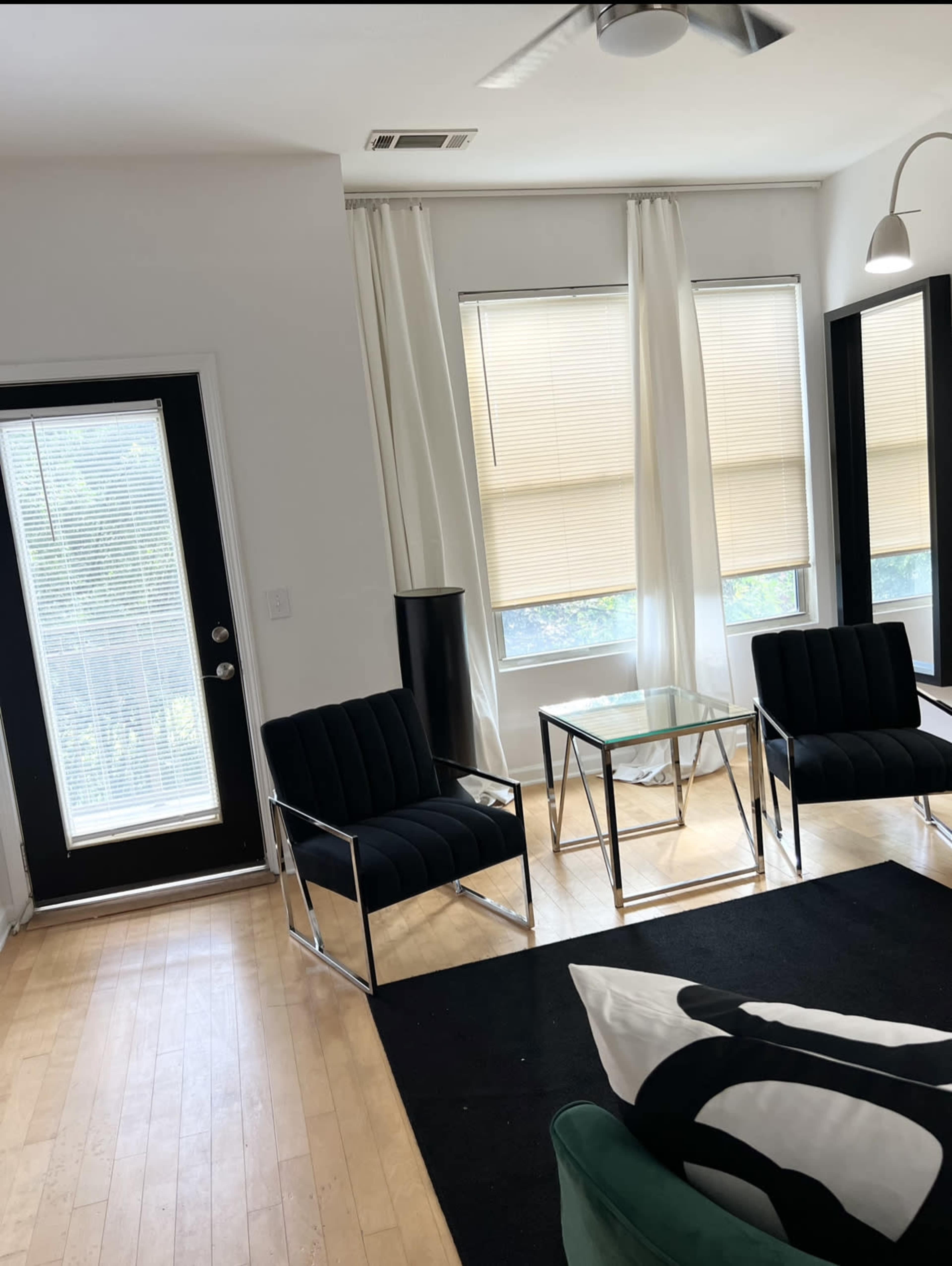 A modern living room with two black chairs, a glass coffee table, and large windows with light-colored shades.