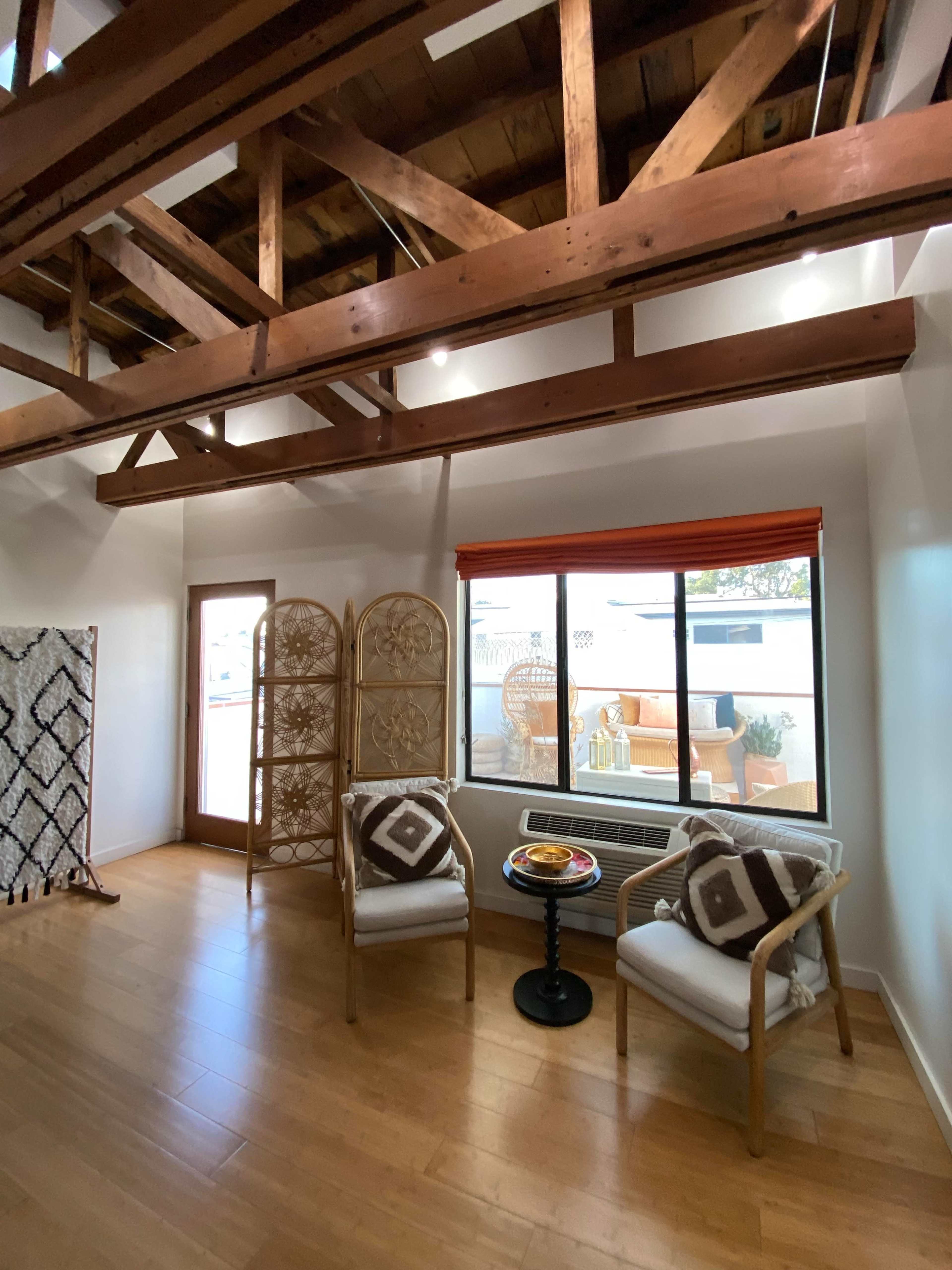 The image shows a spacious room with exposed wooden beams, two rattan chairs, a small side table with a plate of snacks, and a large window overlooking a patio area.