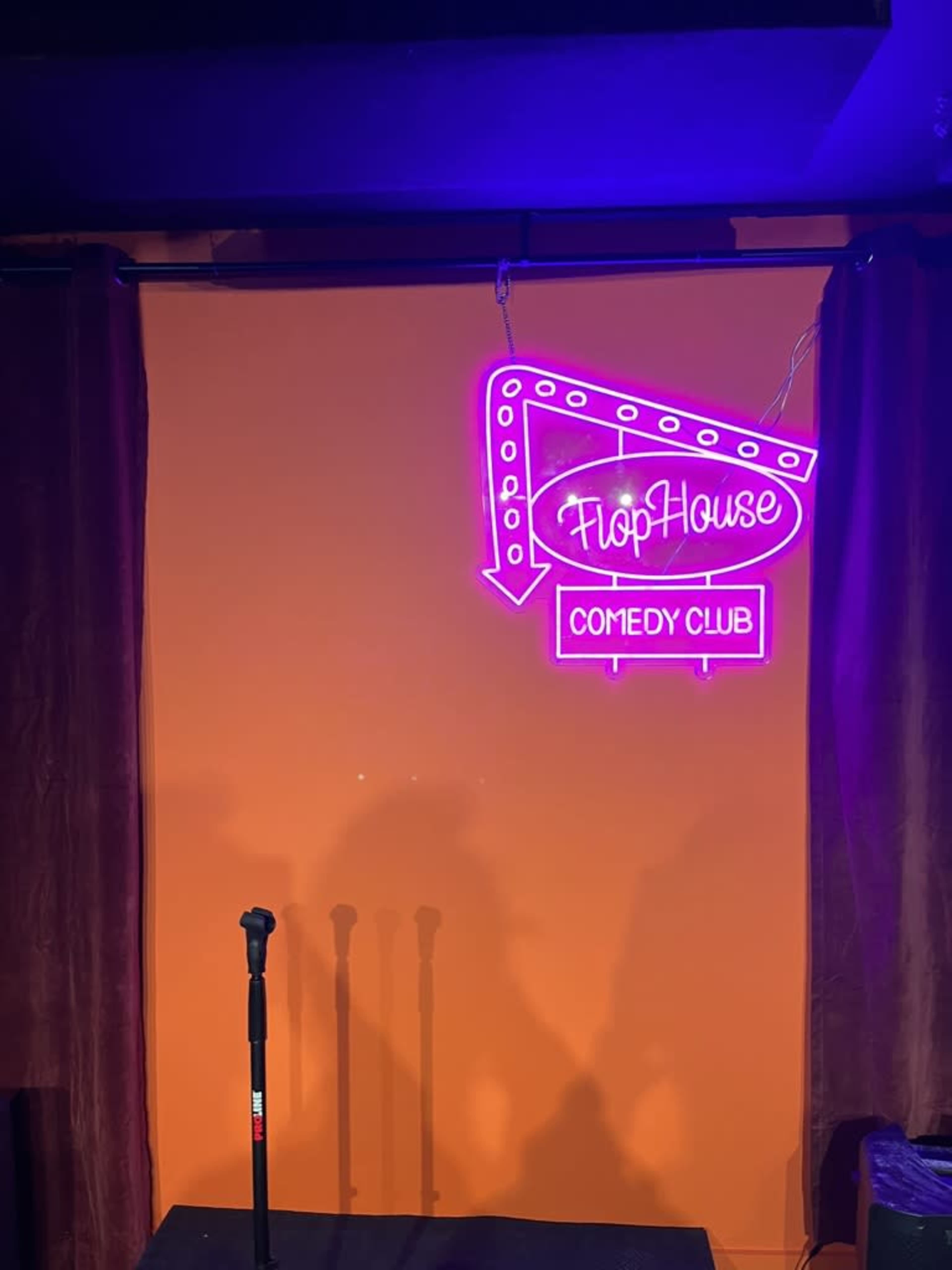 A neon sign for "FlopHouse Comedy Club" illuminated against an orange wall, with a microphone stand in front of it.