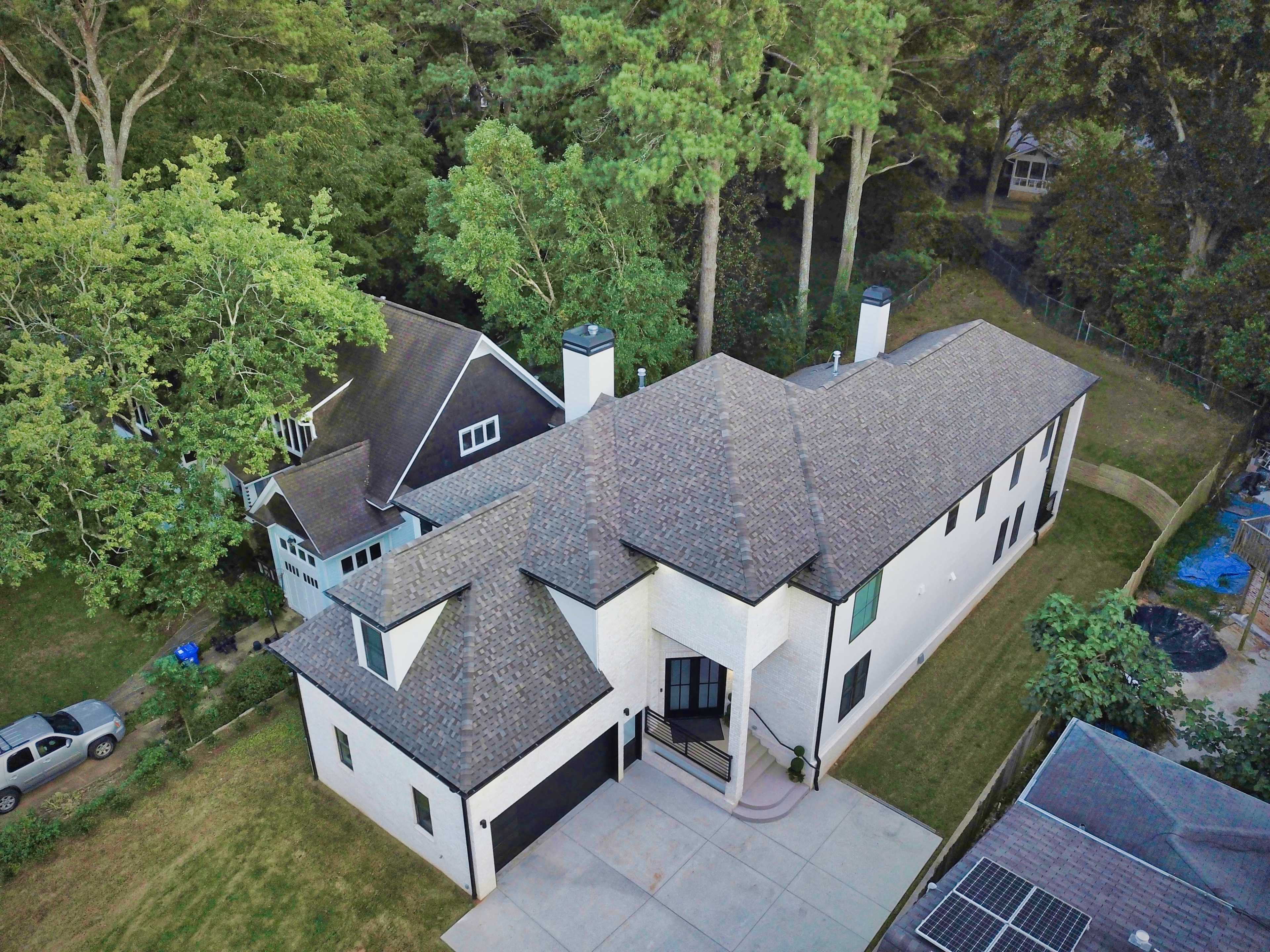 An aerial view shows two houses, one modern and white with a large driveway, and another dark-colored house nestled among trees.