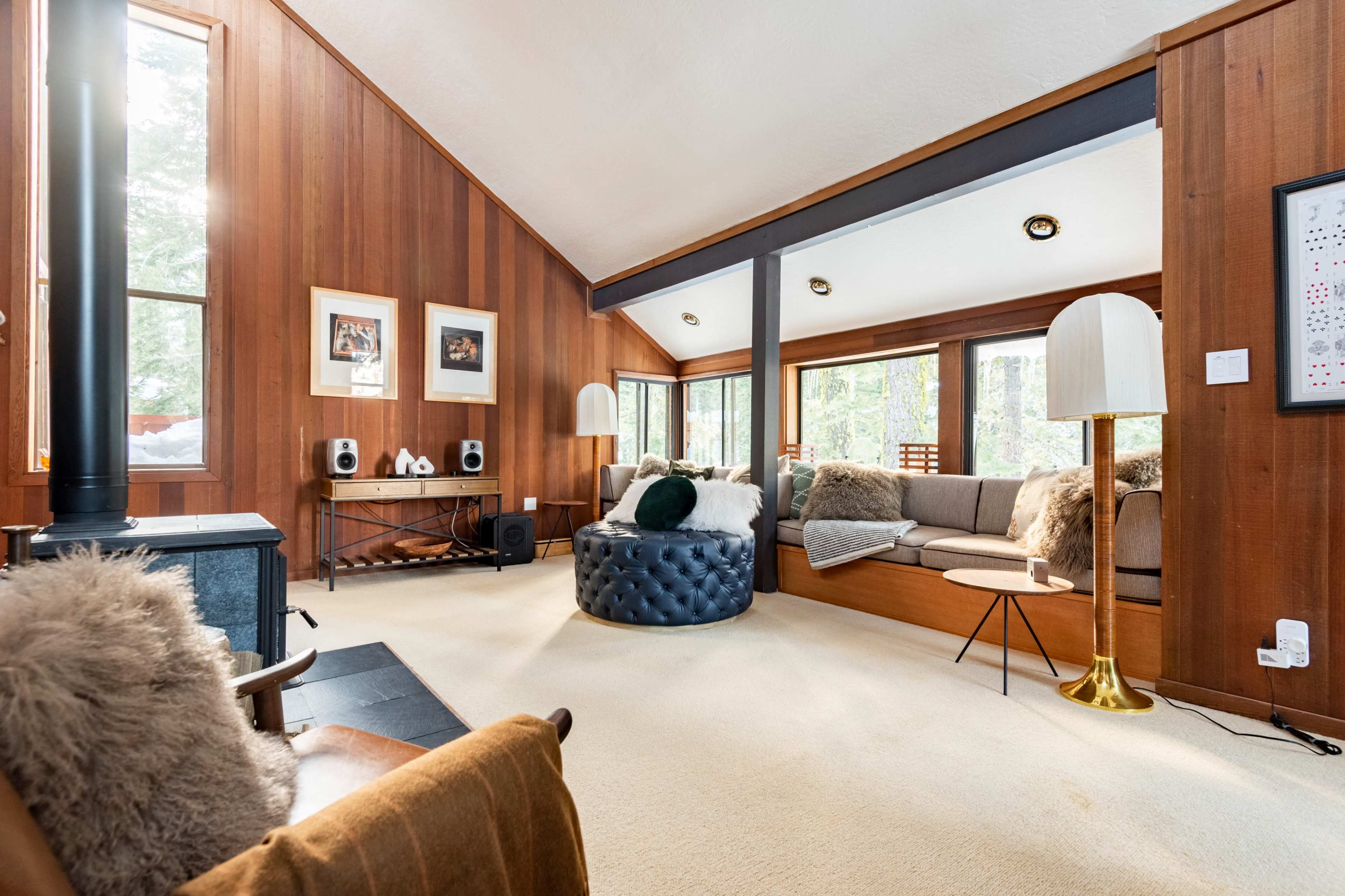 The living room features wooden walls, large windows, a black tufted ottoman, and reupholstered seating arranged around a central stove.