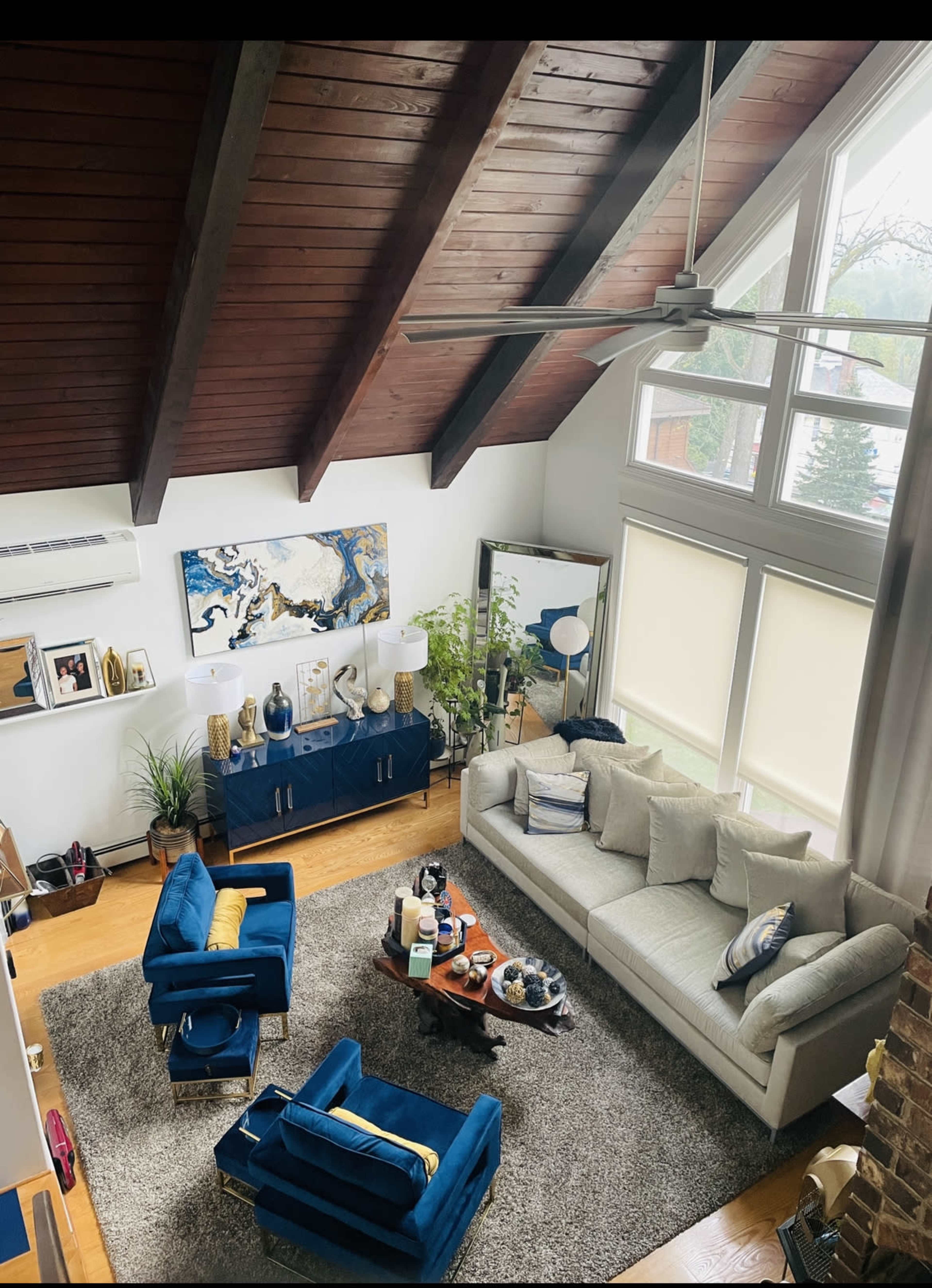 A spacious living room with high wooden ceilings, featuring a large gray sectional sofa, blue accent chairs, a coffee table with decor items, and large windows allowing natural light.