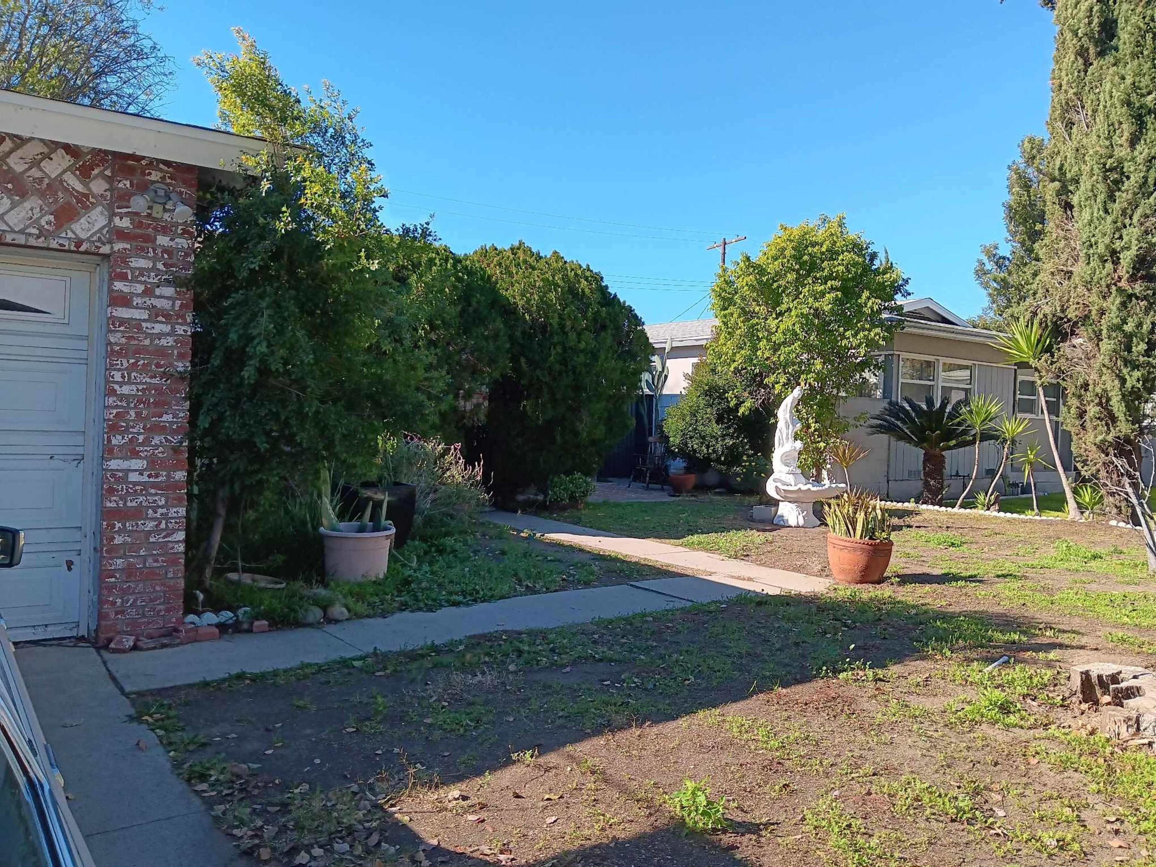 The image shows a garden pathway bordered by shrubs and potted plants, leading towards a house in the background with a statue visible among the greenery.