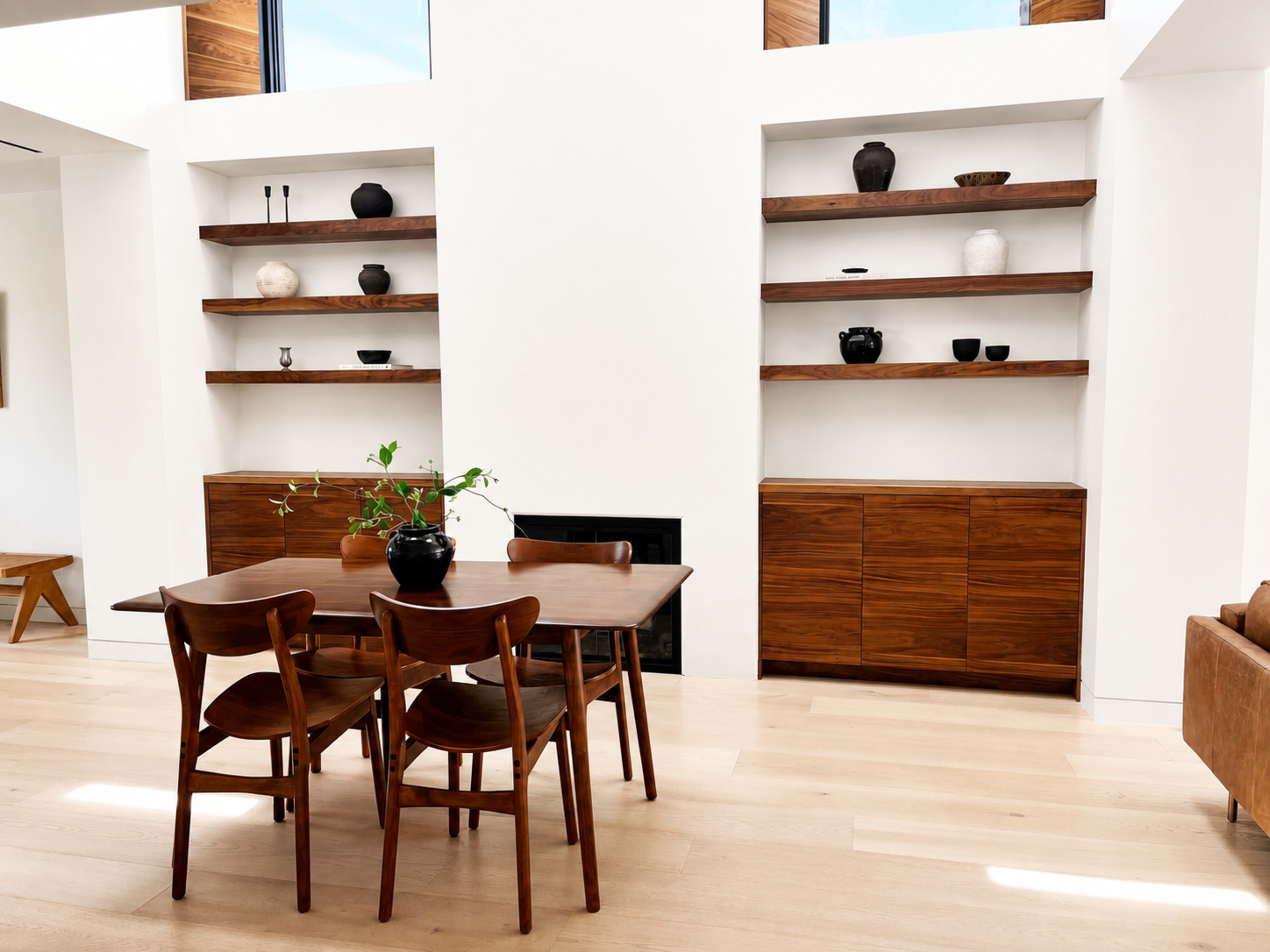 The image shows a modern interior featuring a wooden dining table with four chairs, a wall-mounted shelving unit displaying various objects, and a side cabinet near a small fireplace.