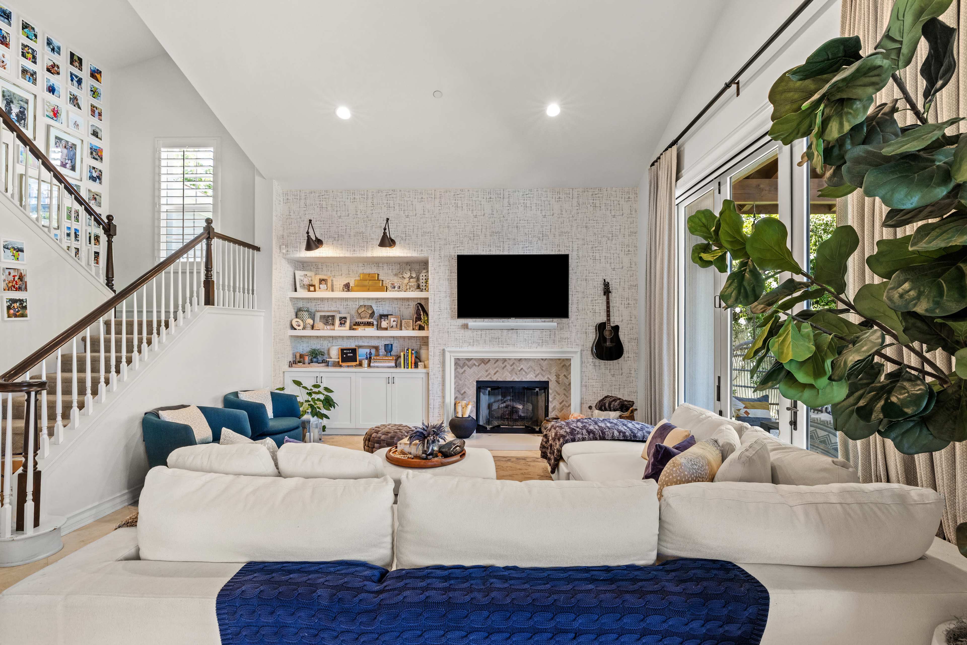 A well-lit living room features a large sectional sofa, a fireplace with a decorative surround, and a mounted television above the mantle, with a staircase and a gallery wall in the background.